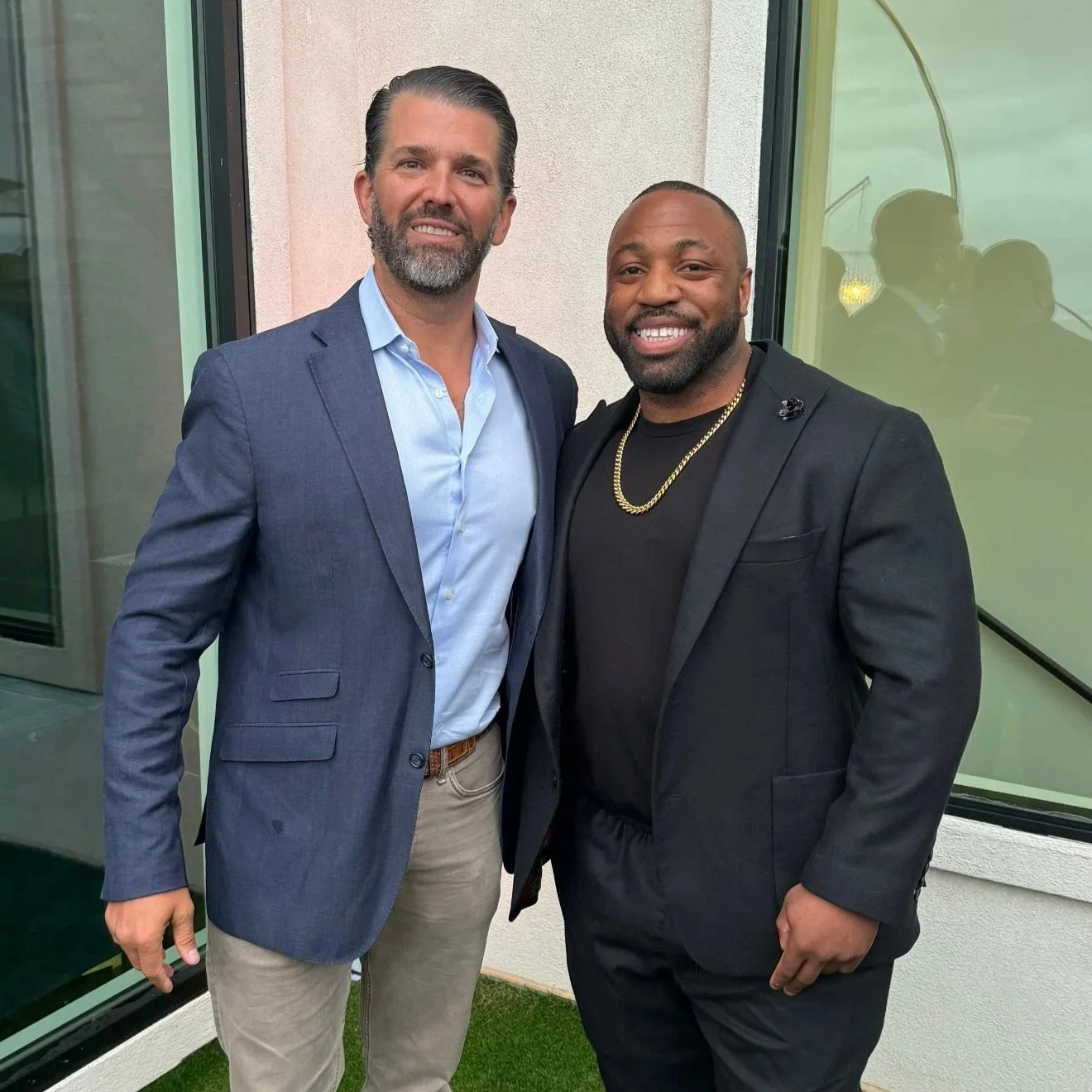 Two men standing outdoors, wearing suits and smiling at the camera.