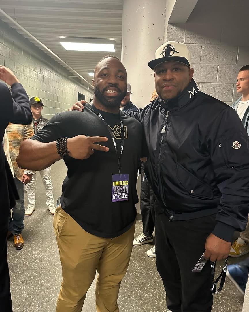Two men posing for a photo at an event, with one showing a peace sign and wearing a black shirt with a speaker guest badge, and the other wearing a black Moncler jacket, a baseball cap, and holding a lanyard, with several people visible in the background in a corridor.