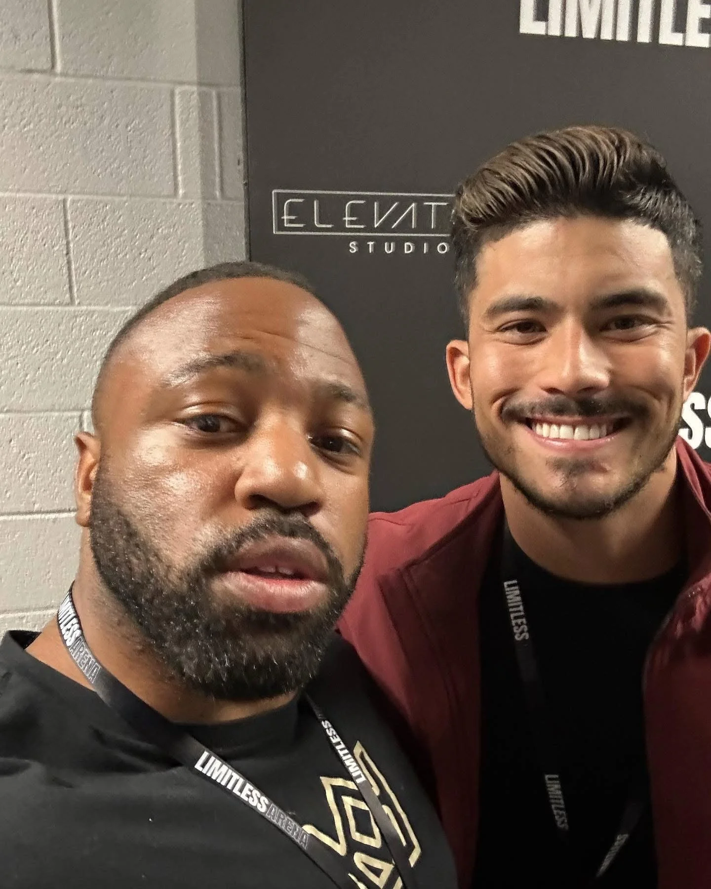 Two men taking a selfie indoors in front of a black wall with white text that reads 'ELEVATE STUDIO'. One man has a beard, is wearing a black shirt and a black lanyard, and is looking at the camera. The other man has short dark hair, is smiling, and is wearing a maroon jacket and a black lanyard.