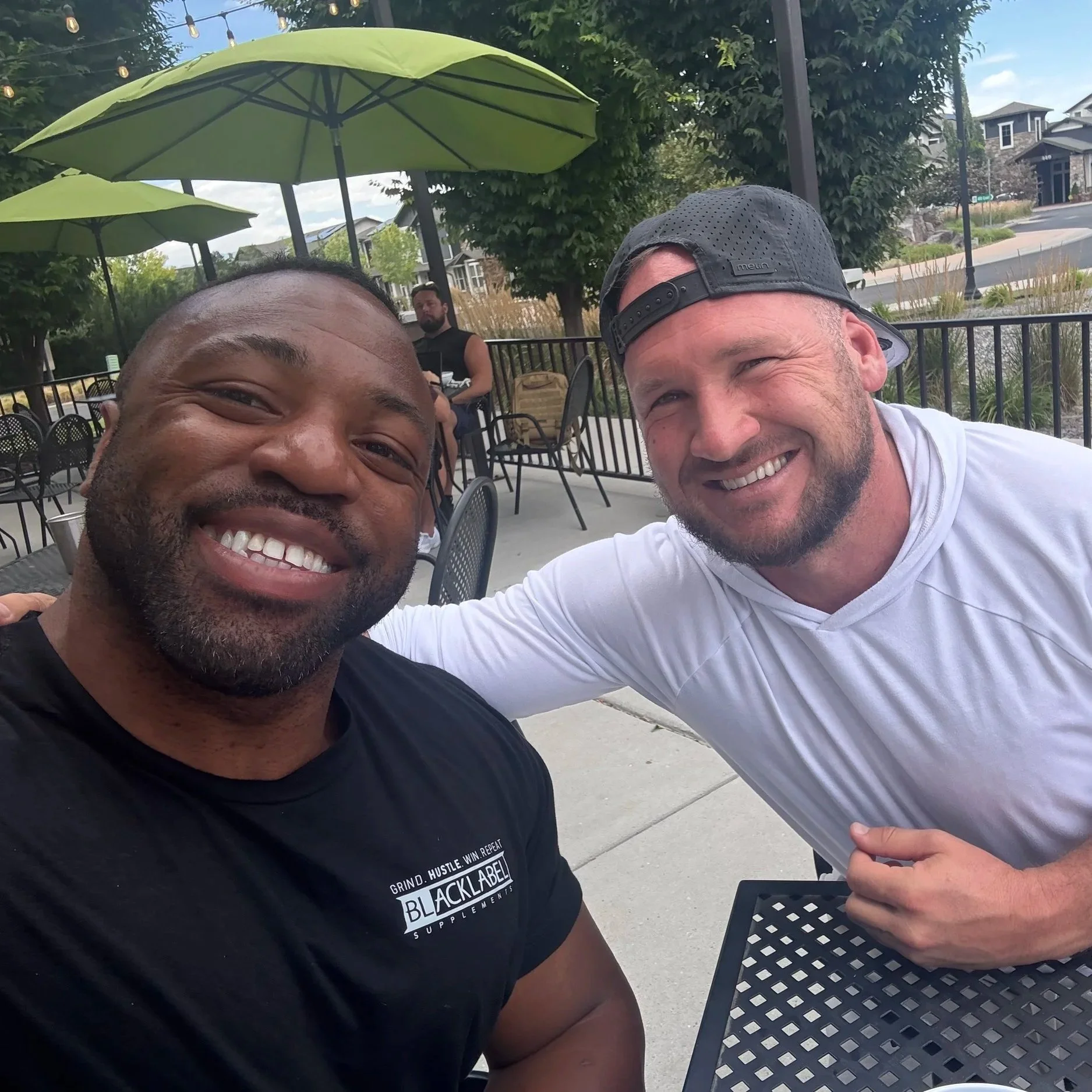 Two men smiling, sitting outdoors at a table with a woman in the background, under green umbrellas.