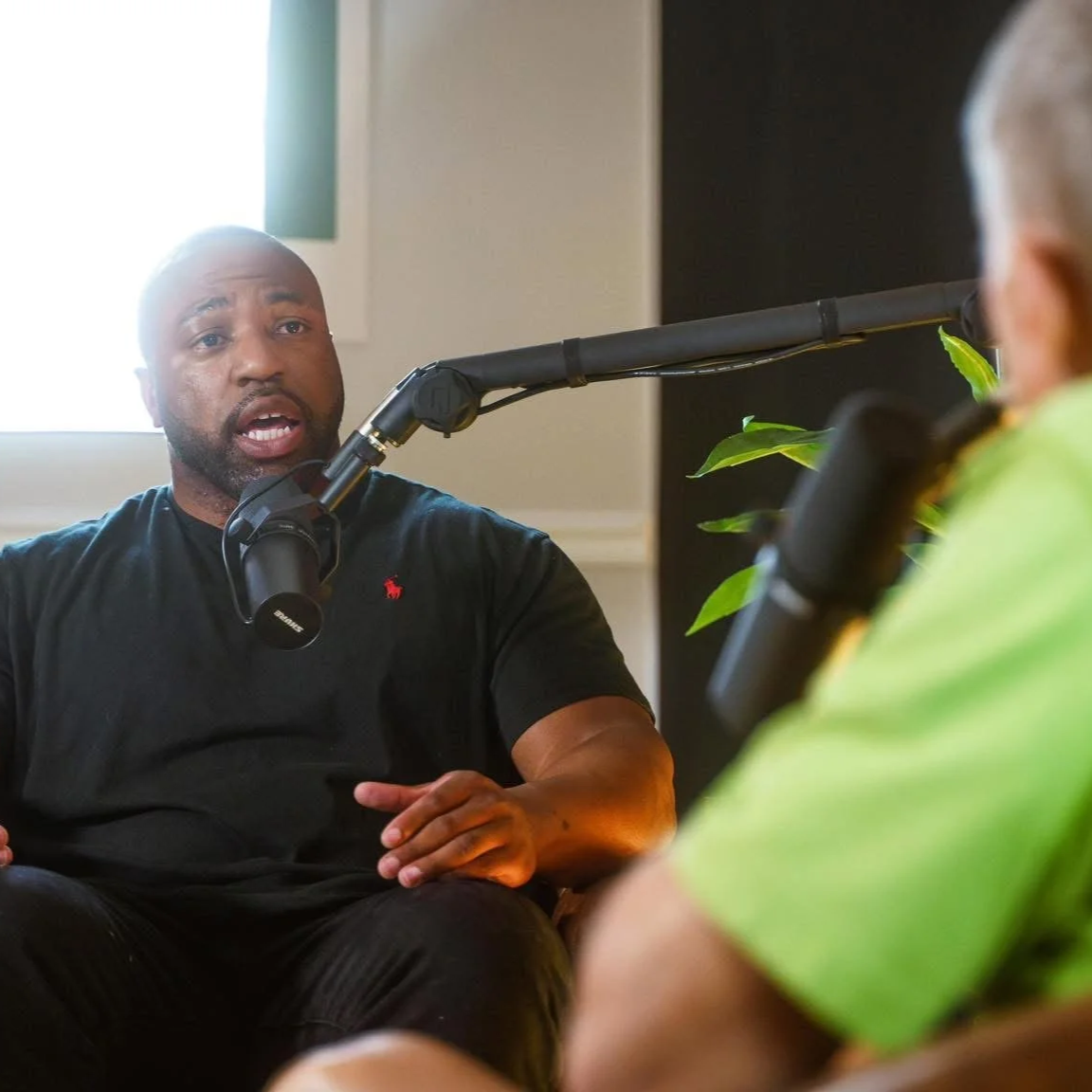 Two men having a conversation in a room, with one man speaking into a microphone, suggesting a podcast or radio interview recording environment.