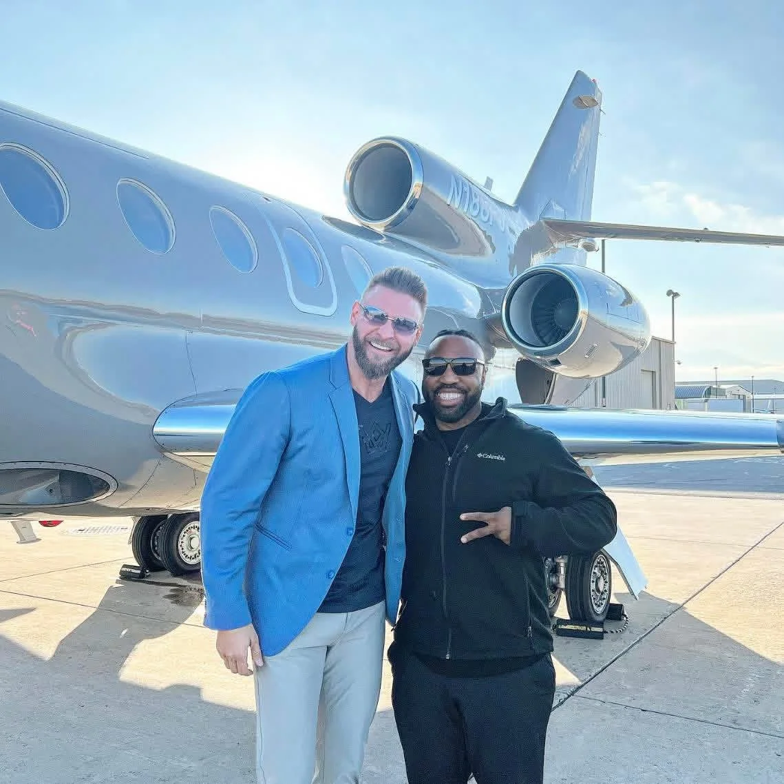Two men smiling and wearing sunglasses in front of a private jet on an airport tarmac during daytime.