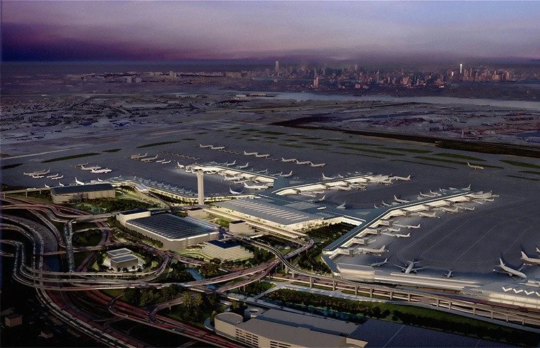 Aerial view of an airport with numerous airplanes parked at gates, surrounded by roads and runways, with a city skyline in the background during twilight.