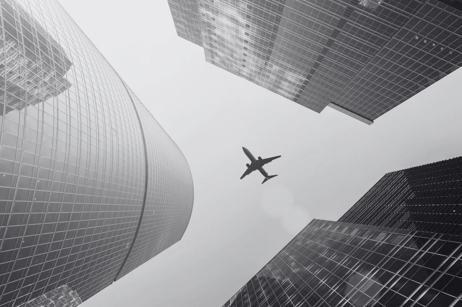 Looking up at a plane flying among tall modern office buildings on a cloudy day.