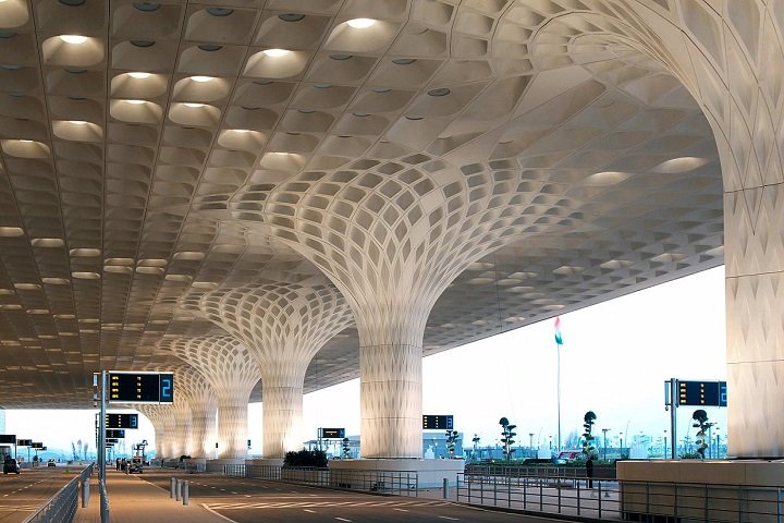 Modern airport terminal with large, sculptural columns and a reflective ceiling, featuring digital information signs and an open pathway outside.