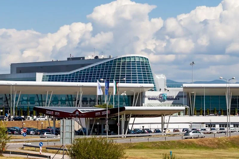 Modern airport terminal building with curved glass facade, security checkpoint, and parked cars in front.