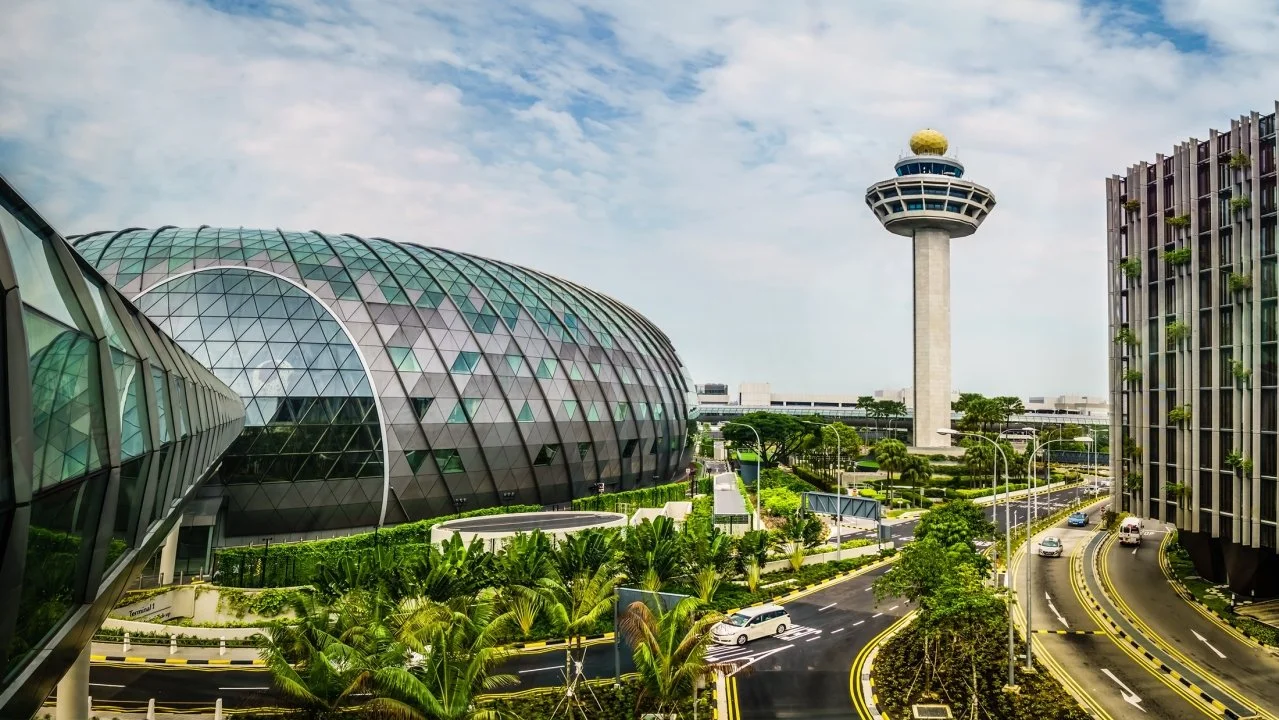 Modern urban scene with futuristic building featuring a curved glass facade, a tall control tower with a round top, and roads with cars surrounded by greenery.