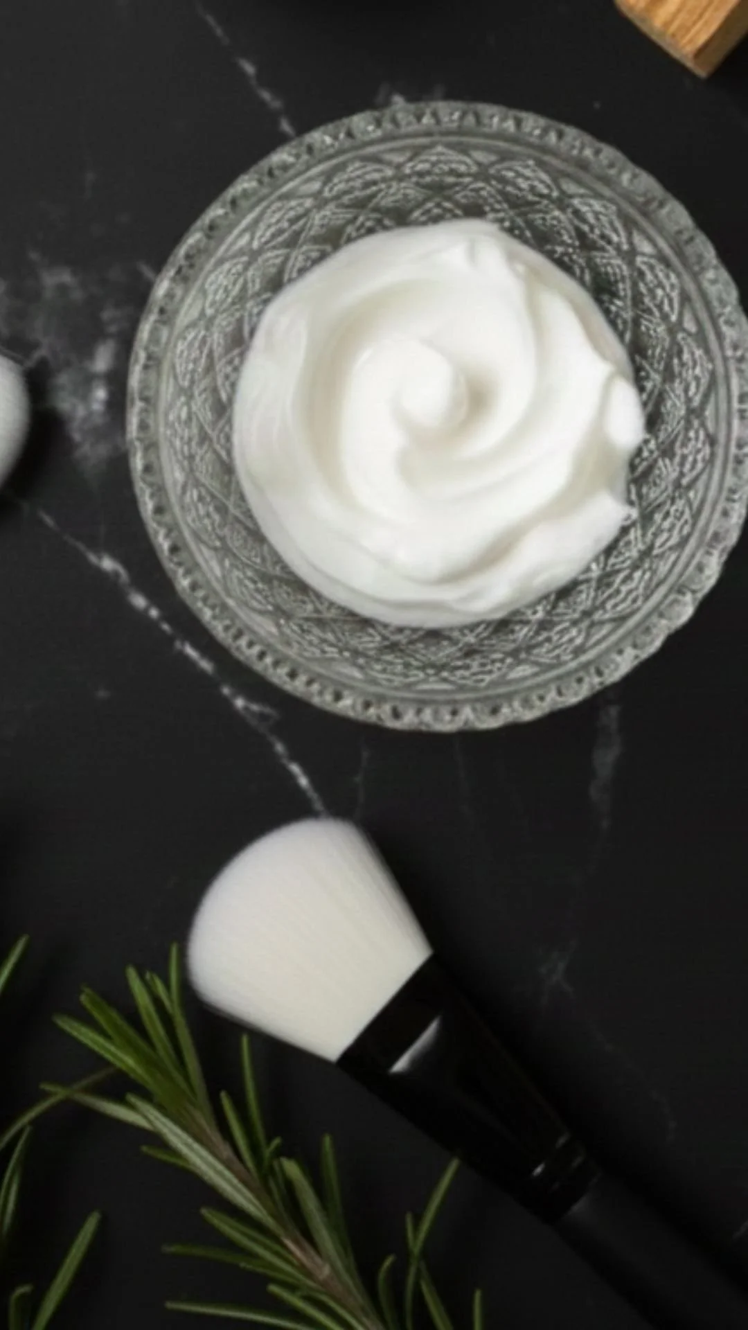 A glass bowl filled with white whipped cream or frosting on a dark surface. Next to it are a makeup brush and a sprig of pine or fir.