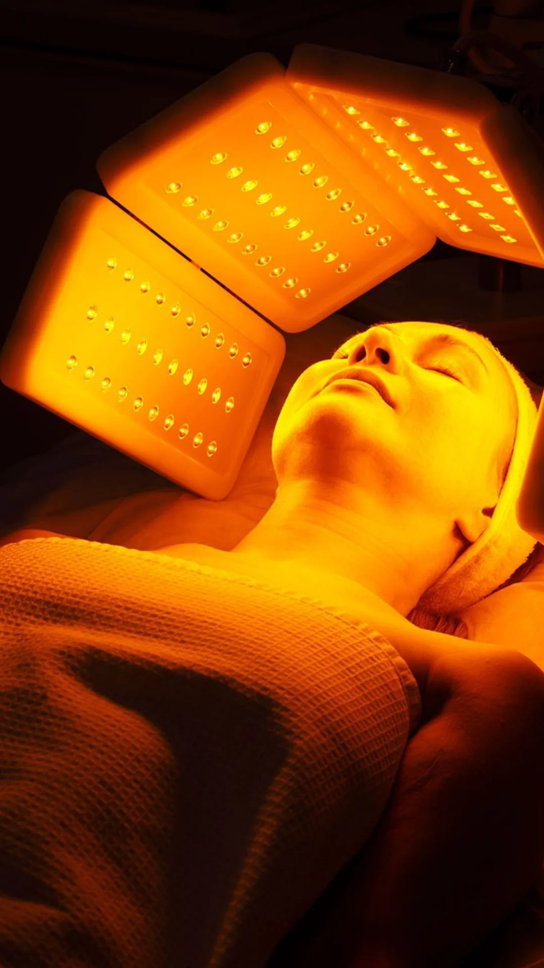 A person lying down with eyes closed, receiving LED light therapy from two orange LED panels above them.