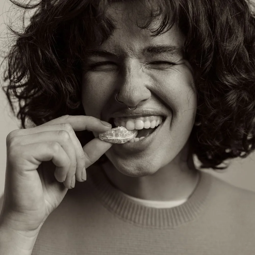 Black and white image of a woman with curly hair and a nose ring eating an orange