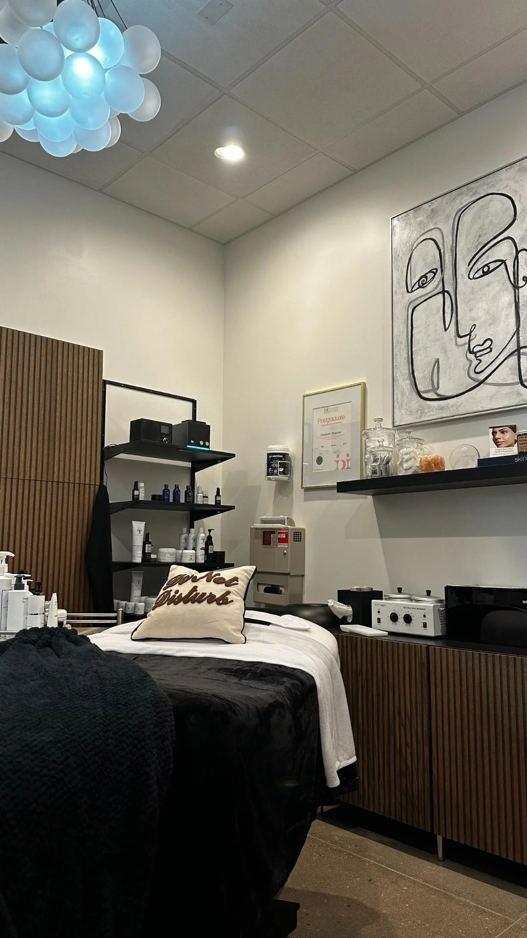 Inside Preparations Skin Spa - Interior of our treatment room with a treatment bed, shelves with skincare products, abstract wall art, and ceiling lights with balloon-like fixtures.