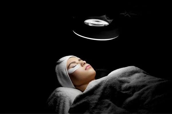 A woman lying on a bed with her eyes closed, covered with a blanket, receiving a facial treatment in a dark spa or clinic setting, with a bright light above her face.