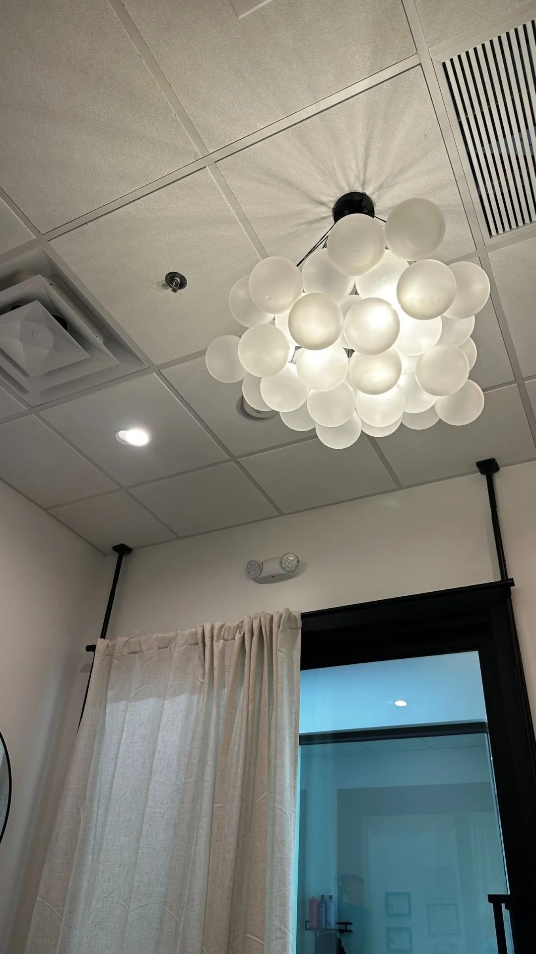 Inside Preparations Skin Spa - Ceiling light fixture with multiple white, spherical bulbs in a cluster installation, in a room with white curtains, a glass door, and ceiling tiles.