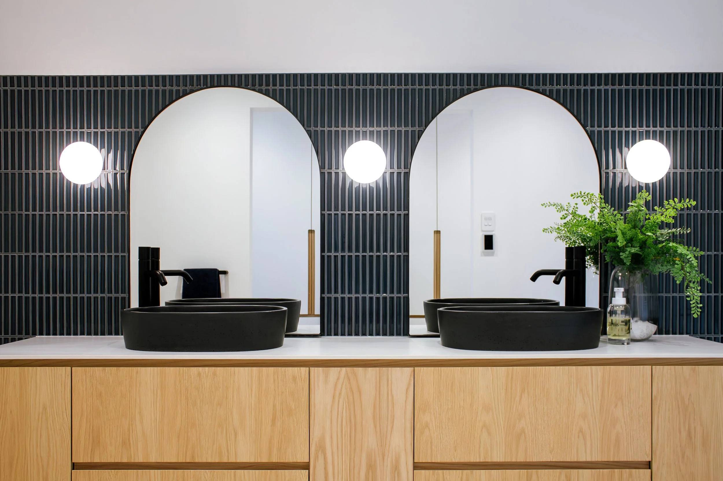 Modern bathroom vanity with two black vessel sinks, black faucets, and large mirrors with black grid frames. There are two round wall sconces, a green potted fern, and a soap dispenser on the countertop. Wooden cabinetry below.