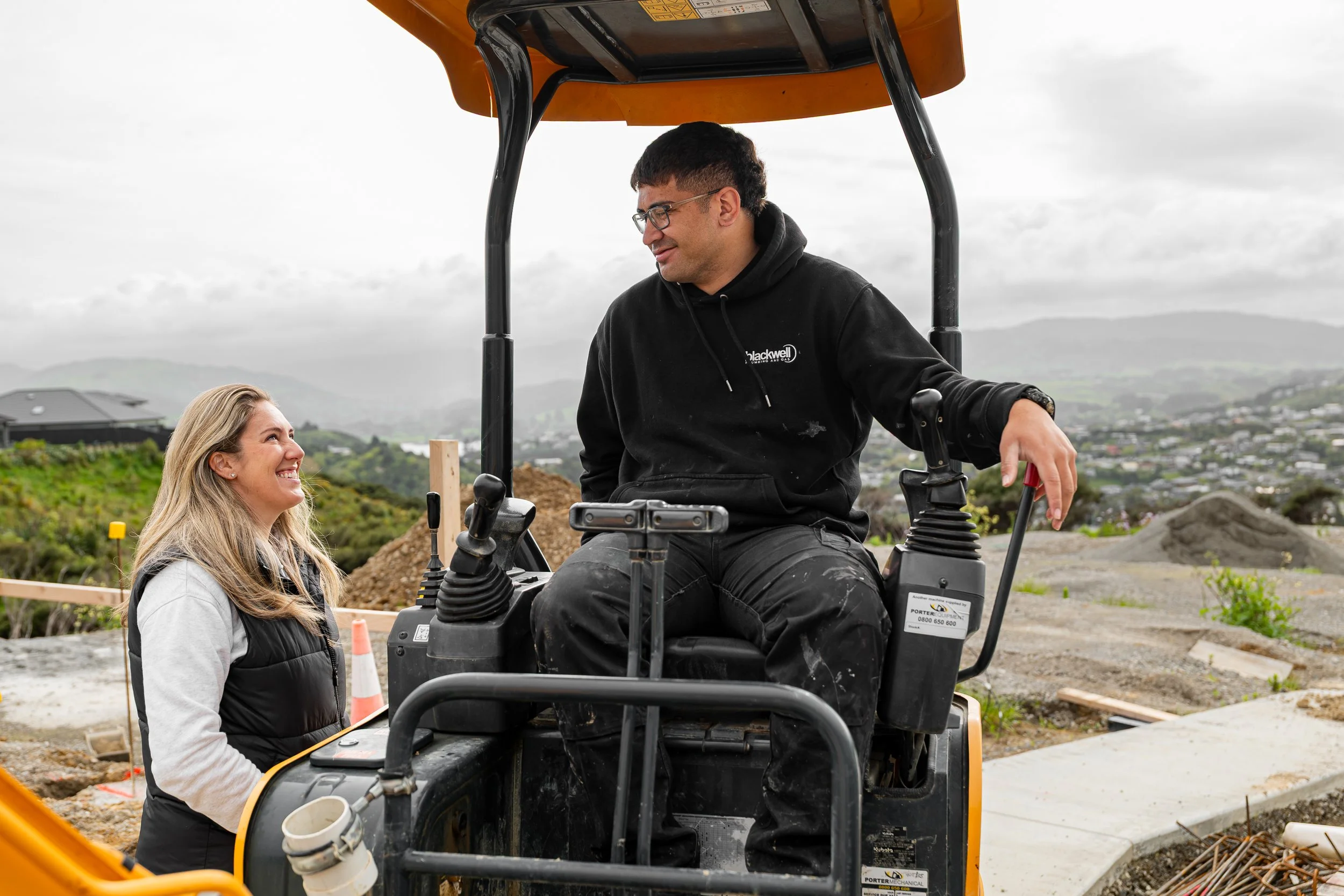 A man on a construction vehicle talking to a woman at a construction site, with hills and houses in the background.