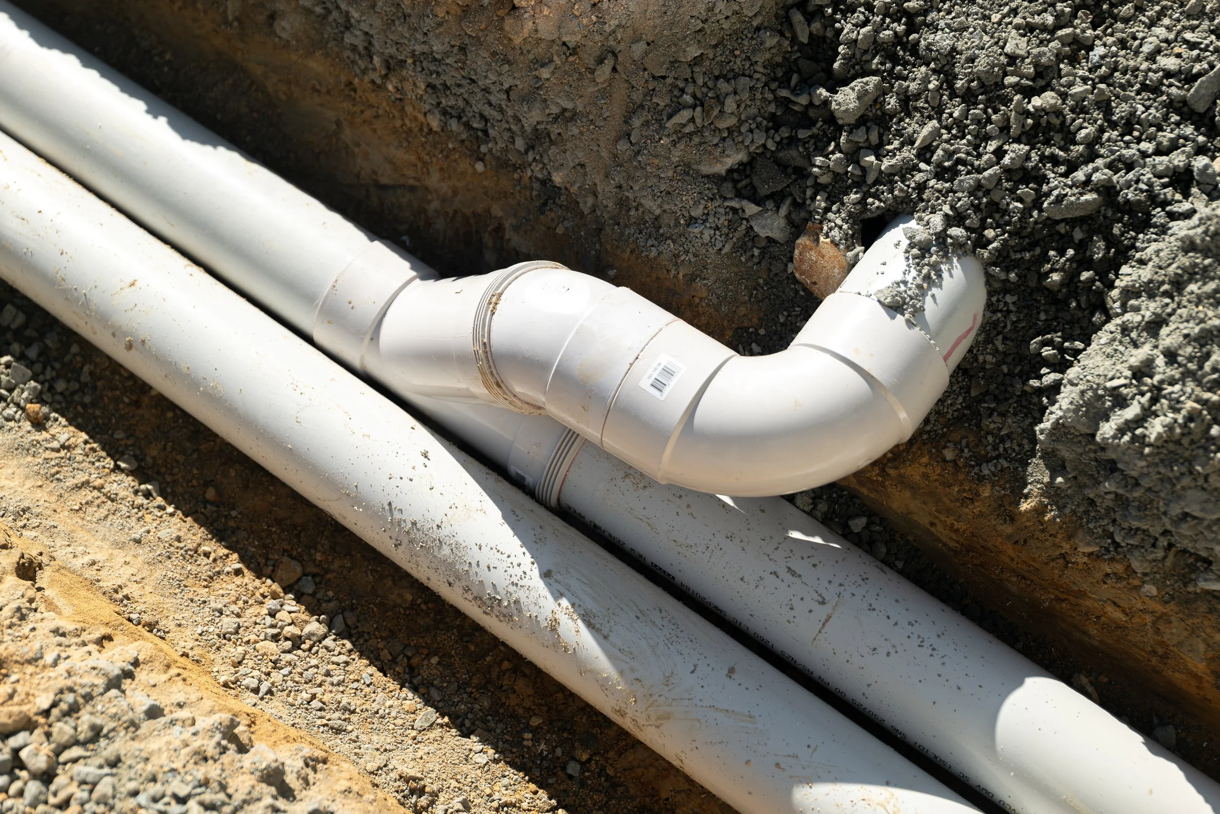 White PVC pipes installed underground with a trench filled with dirt and gravel around them.