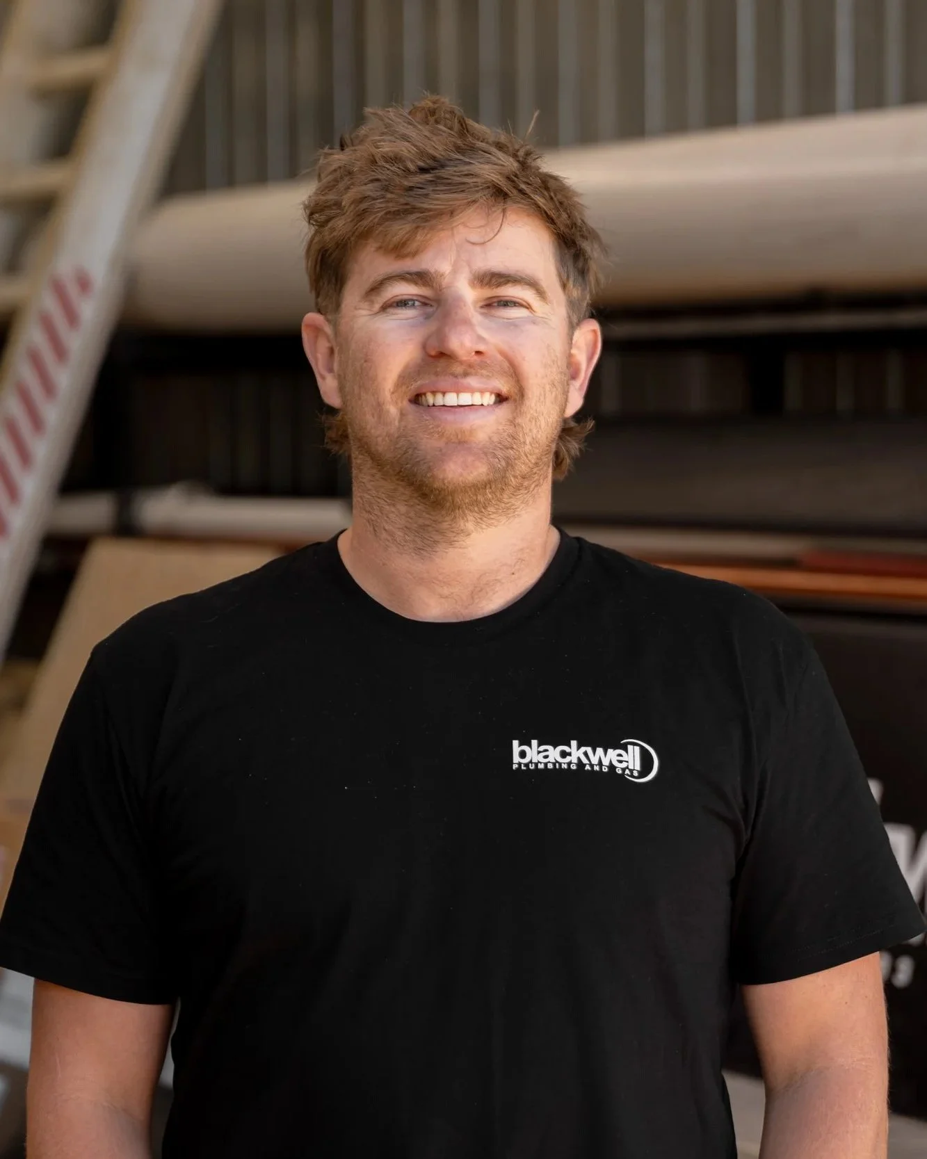 A photo of Josh Blackwell. With brown hair and a beard wearing a black T-shirt with the logo "Blackwell Plumbing and Gas" stands outdoors with what appears to be a building or storage area in the background.