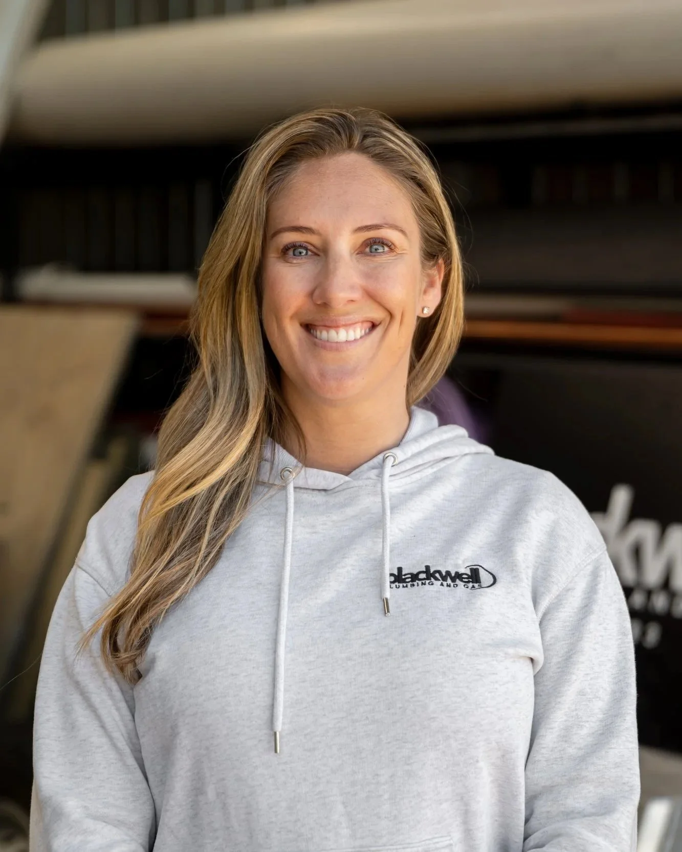 A photo of Amy Blackwell. With long blonde hair smiling, wearing a gray hoodie with a logo that reads "Blackwell Plumbing and Gas", standing outdoors with a blurred background.