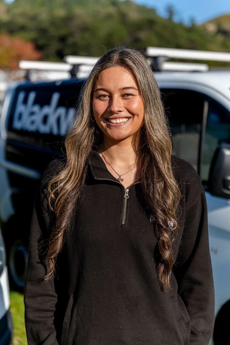 A photo of Victoria Stowers, smiling outdoors in front of a vehicle with a ladder on top on a sunny day.