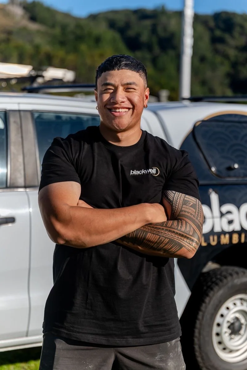 A smiling man with short black hair and a tattoo on his left arm stands with crossed arms in front of a construction site, wearing an orange safety vest over a black shirt.