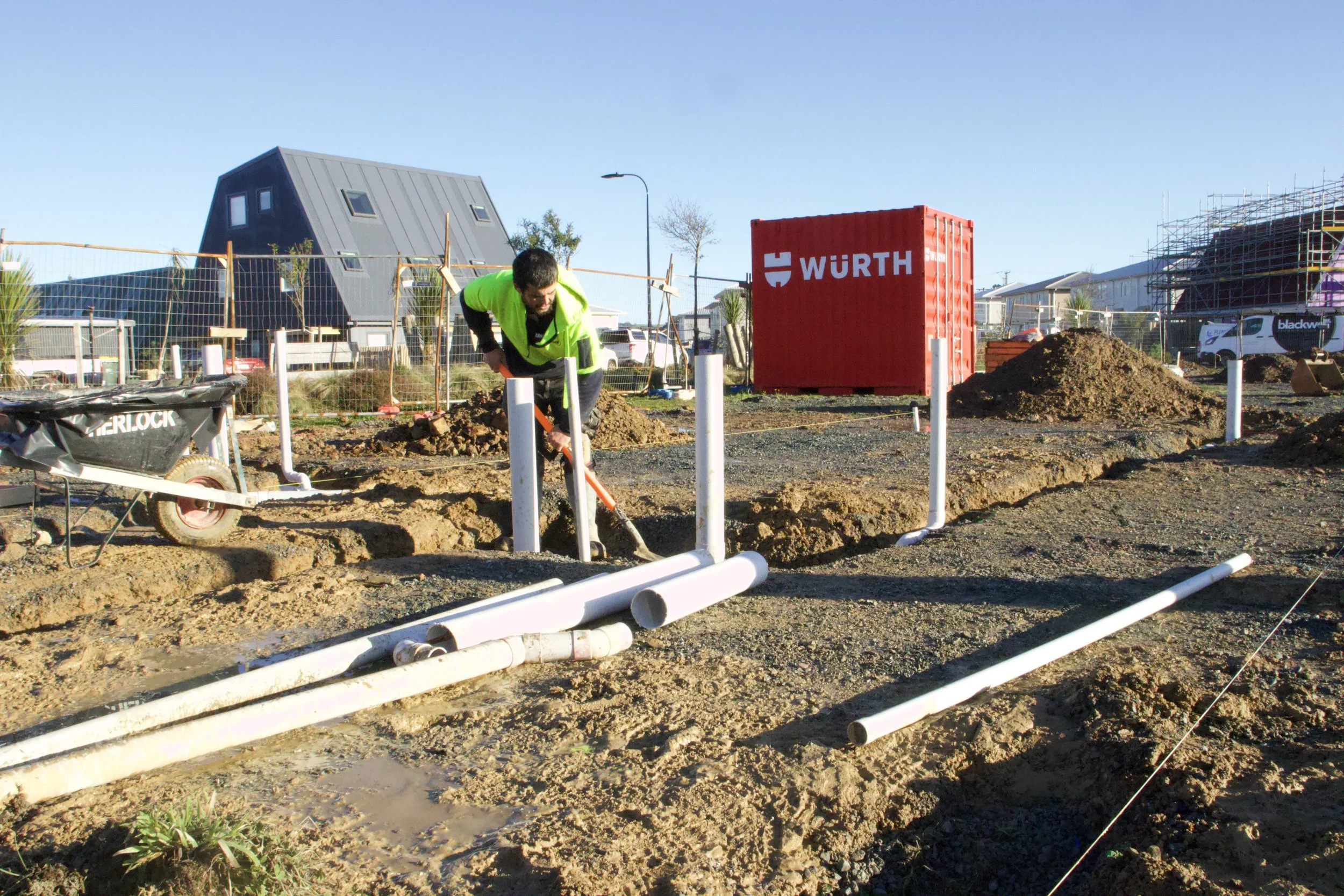 Construction worker in a yellow safety vest working on laying pipes at a construction site with buildings in the background.
