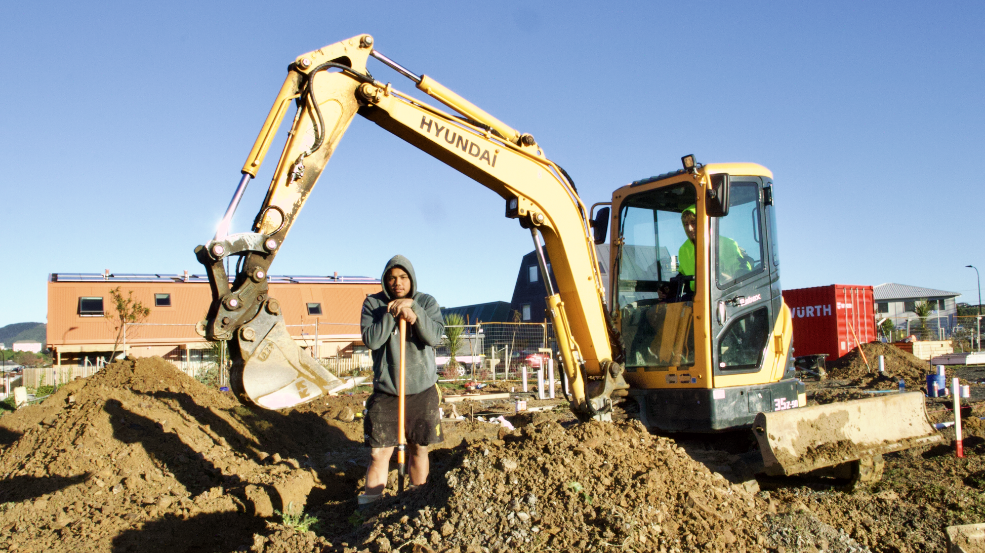Two construction workers at a construction site, one in a gray hoodie and shorts holding a shovel, the other in a high visibility shirt operating a yellow Hyundai mini excavator, blue sky in the background.