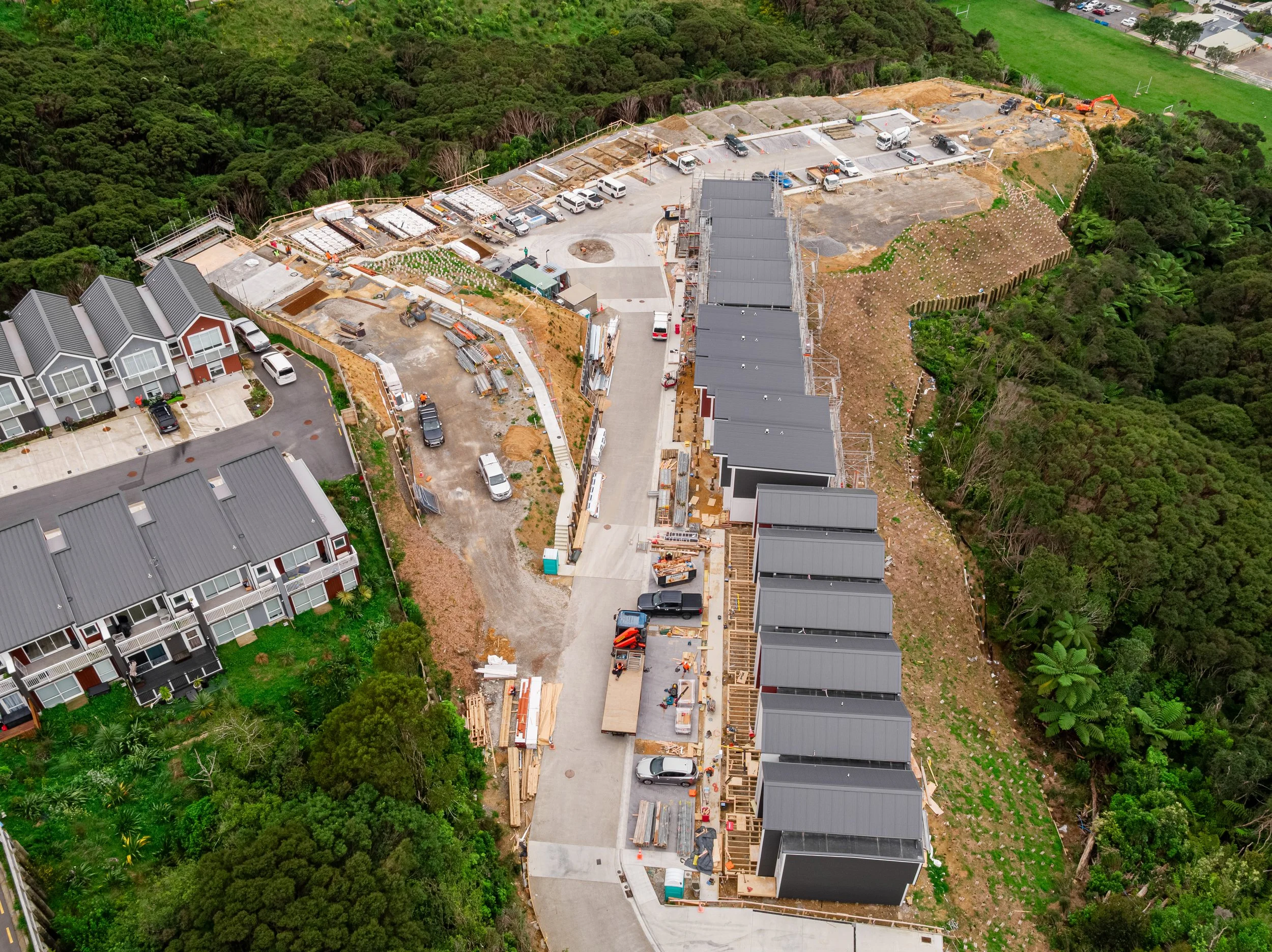 Aerial view of a housing construction site with partially built townhouses, construction materials, and parked trucks, situated next to a residential area with new townhomes and surrounded by green trees.