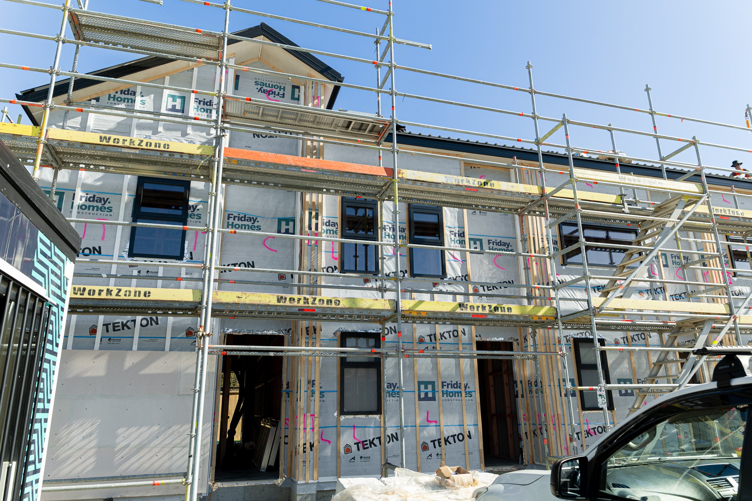 A house under construction with scaffolding, windows installed, and building materials around. The house is wrapped with weatherproofing material branded 'Friday Homes' and 'Tekton.'