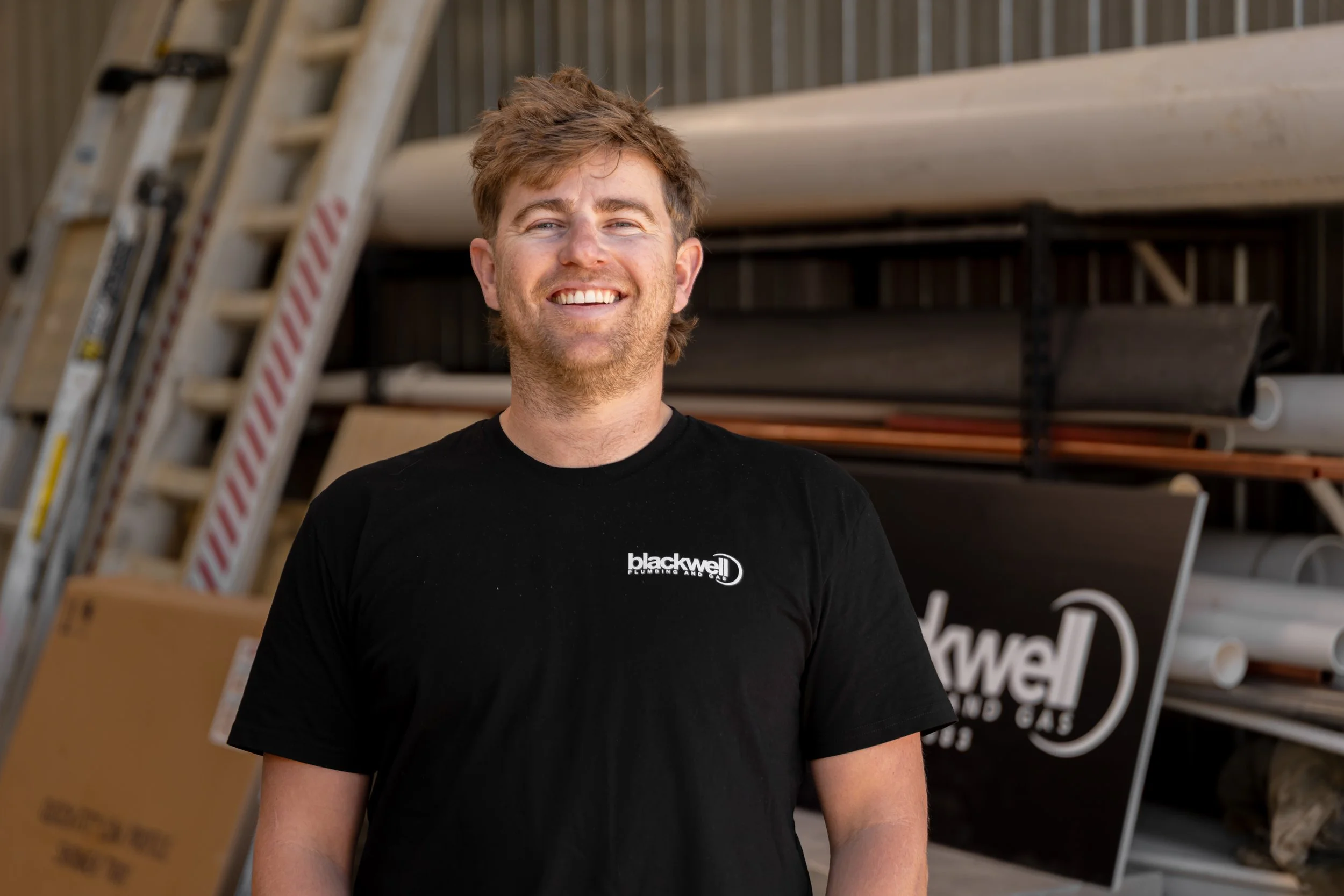 Young man smiling, wearing a black t-shirt with 'blackwell' logo, standing in hardware or plumbing store with pipes and ladders in background.