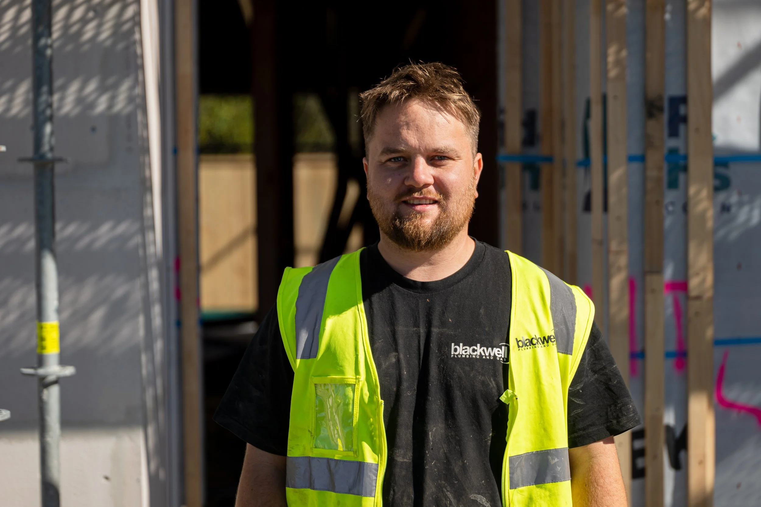 A construction worker wearing a black t-shirt with a company logo and a yellow safety vest standing on a construction site with wooden framing in the background.