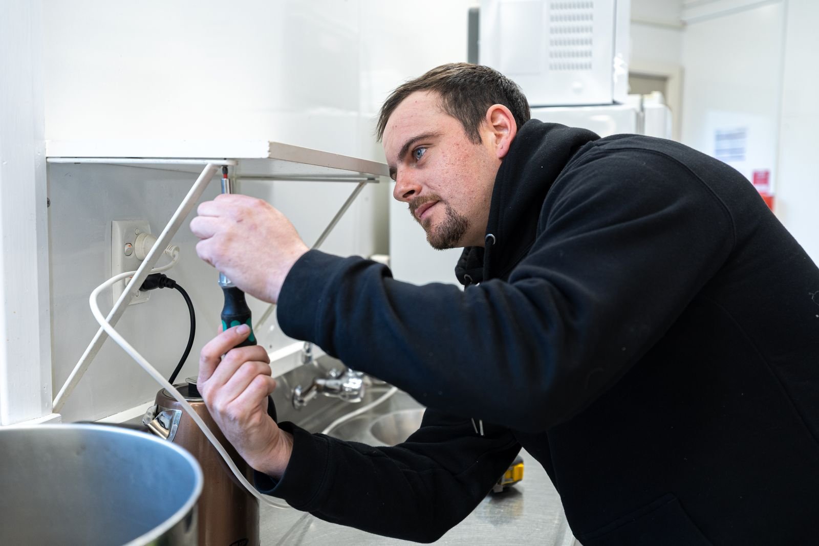 A man in a black hoodie working on an electrical outlet under a kitchen cabinet, using a screwdriver.