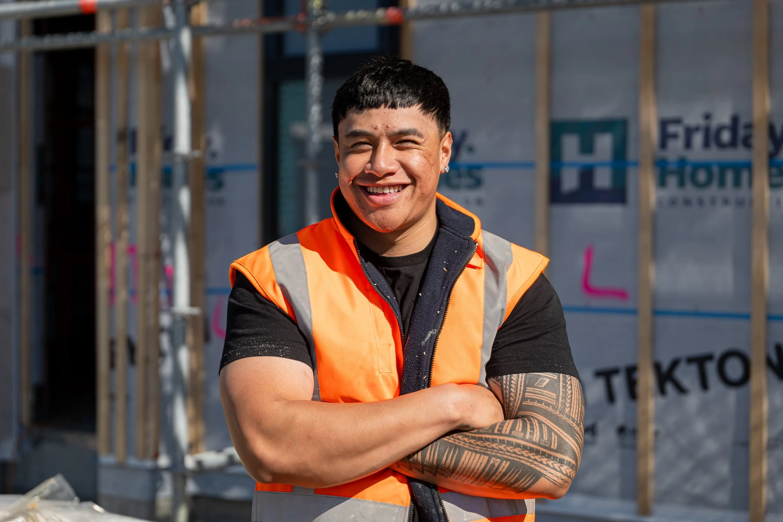 A smiling man with short black hair and a tattoo on his left arm stands with crossed arms in front of a construction site, wearing an orange safety vest over a black shirt.