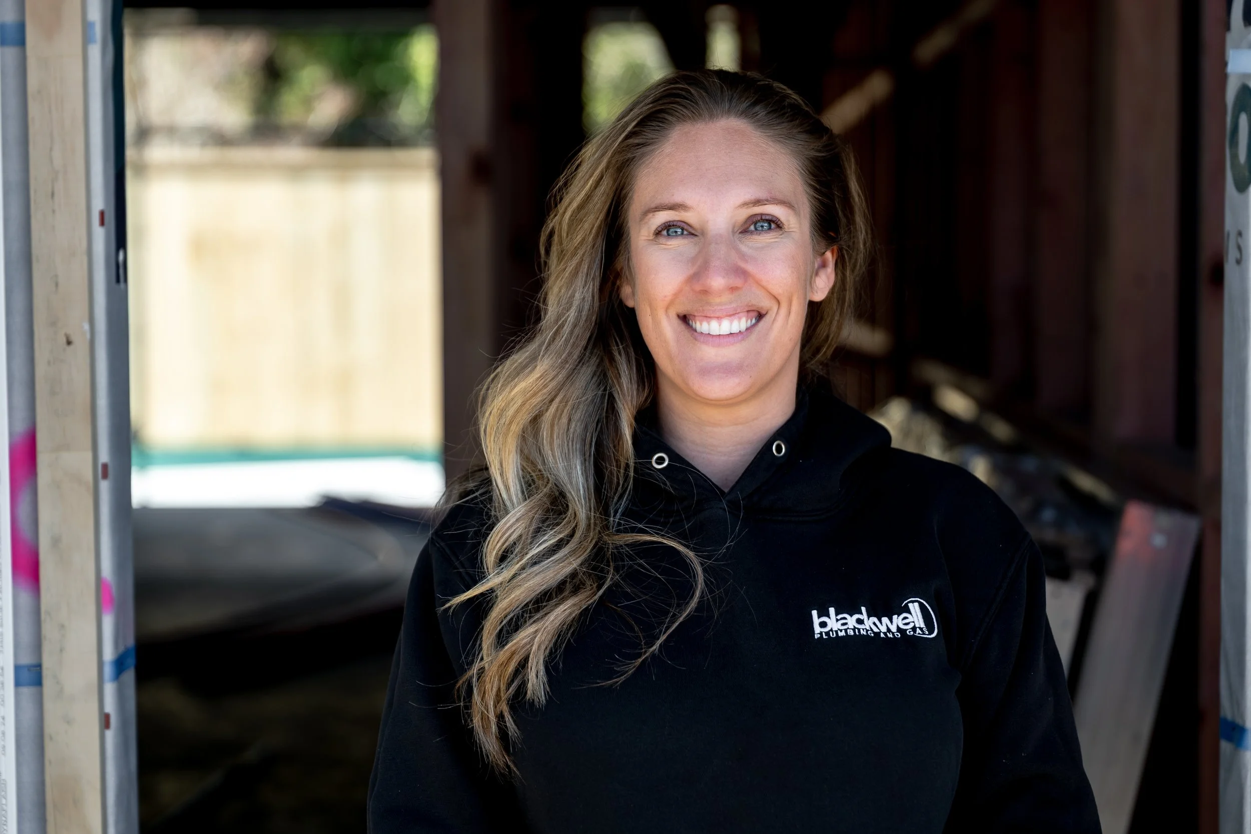 A woman smiling at the camera, wearing a black hoodie with a logo that reads 'Blackwell Plumbing and Gas,' in a partially constructed building with wooden framing.
