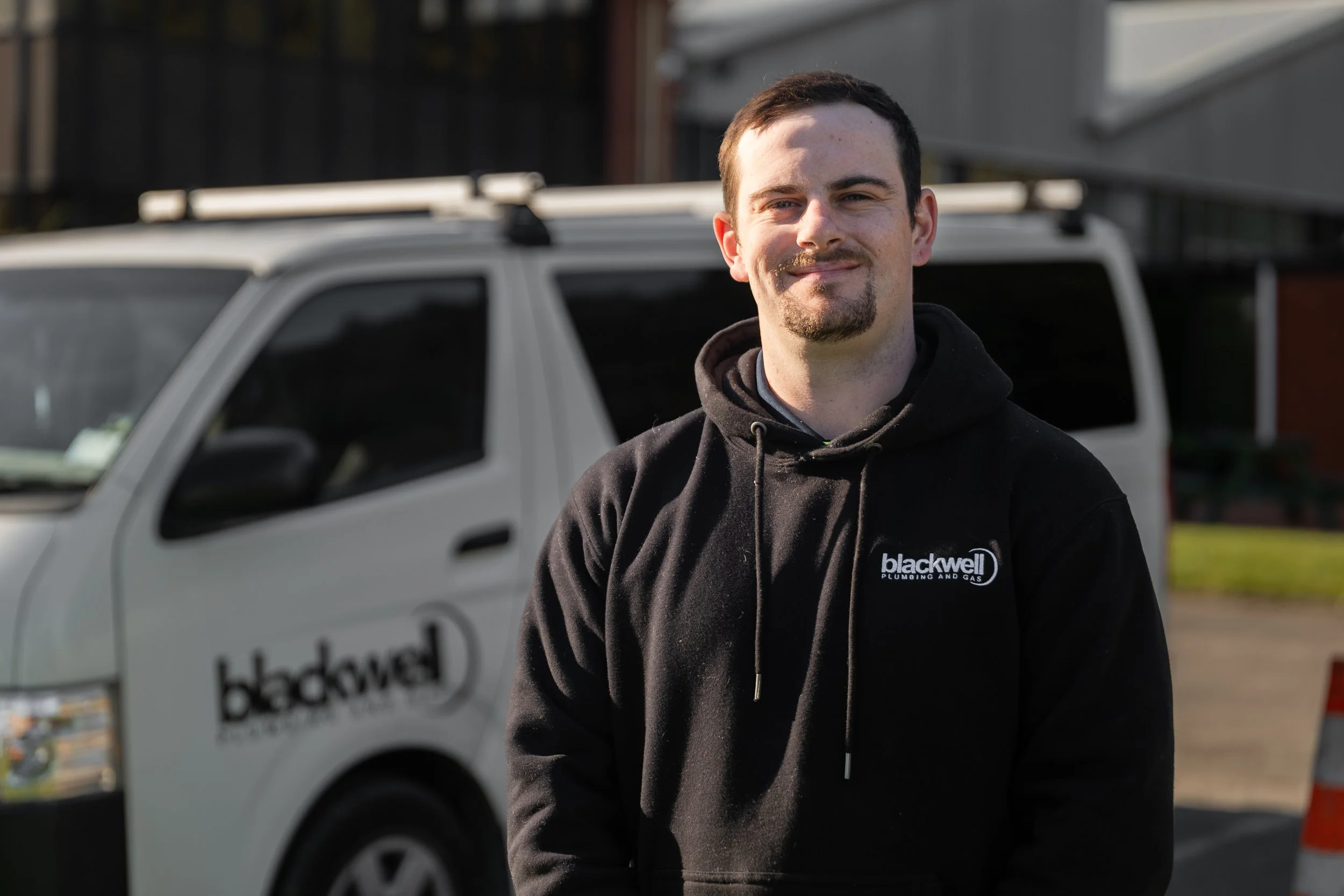 A young man with short hair and a goatee smiling while standing outside in front of a white utility van with black text that reads 'blackwell plumbing and gas.' He is wearing a black hoodie with the same logo and appears to be in a residential or commercial area during daylight.