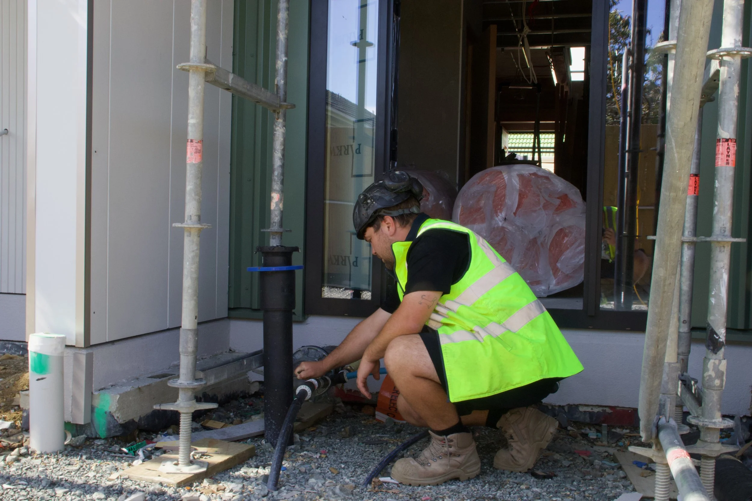 A construction worker wearing a safety vest, helmet, and boots crouches down working on plumbing outside a building under construction. Scaffolding surrounds him, and a large plastic bag filled with building materials is nearby.