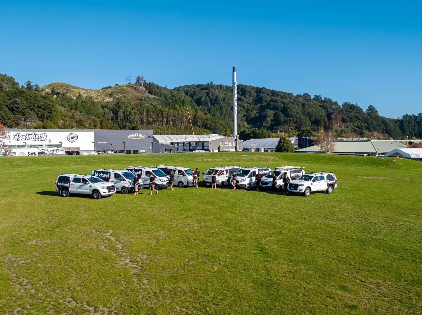 A group of white service vehicles with Blackwells branding parked on a grassy area. Several people are standing near the vehicles, and the background features a building complex with advertising signs and green hills under a clear blue sky.