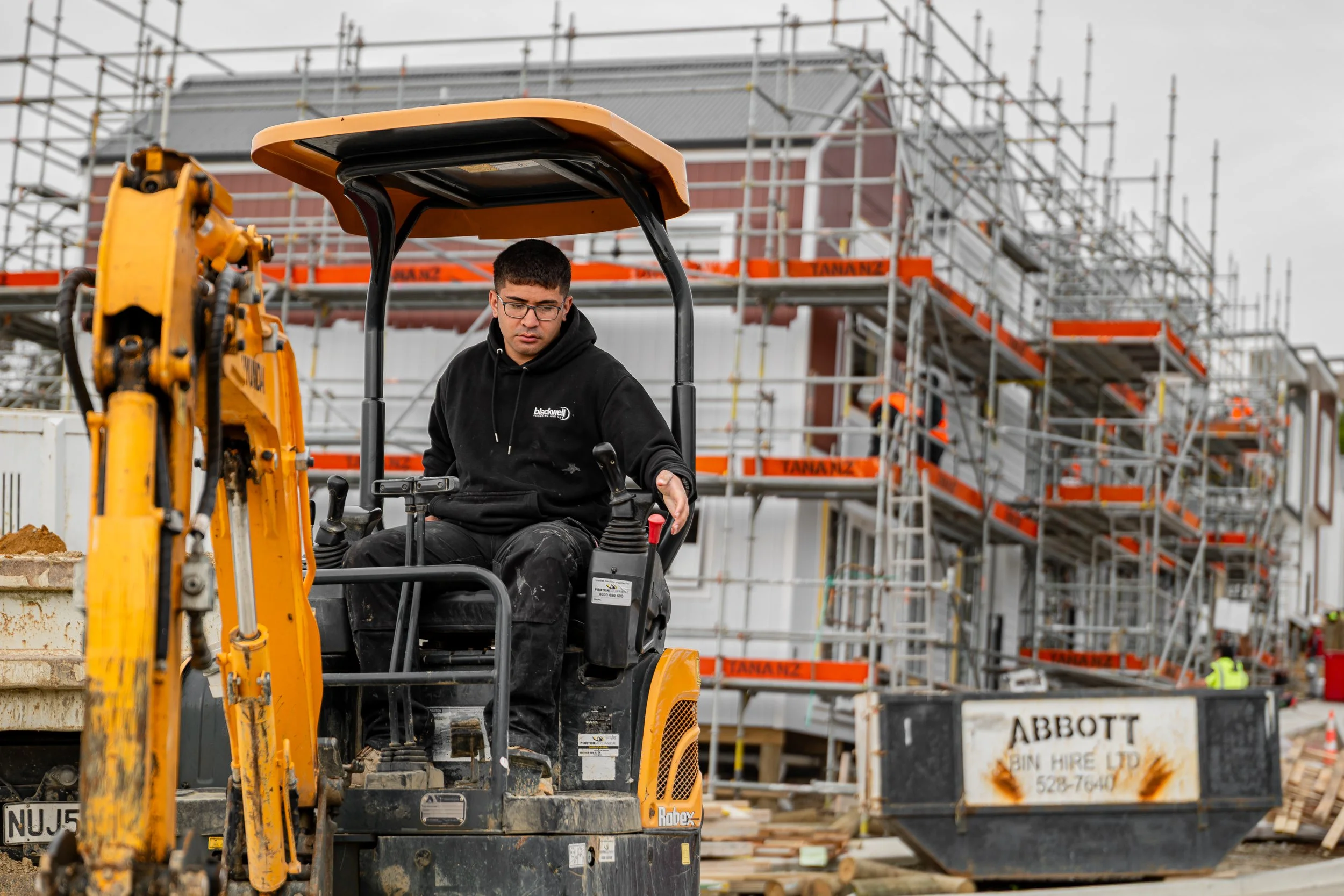 A man operating a small construction vehicle on a building site with scaffolding in the background.