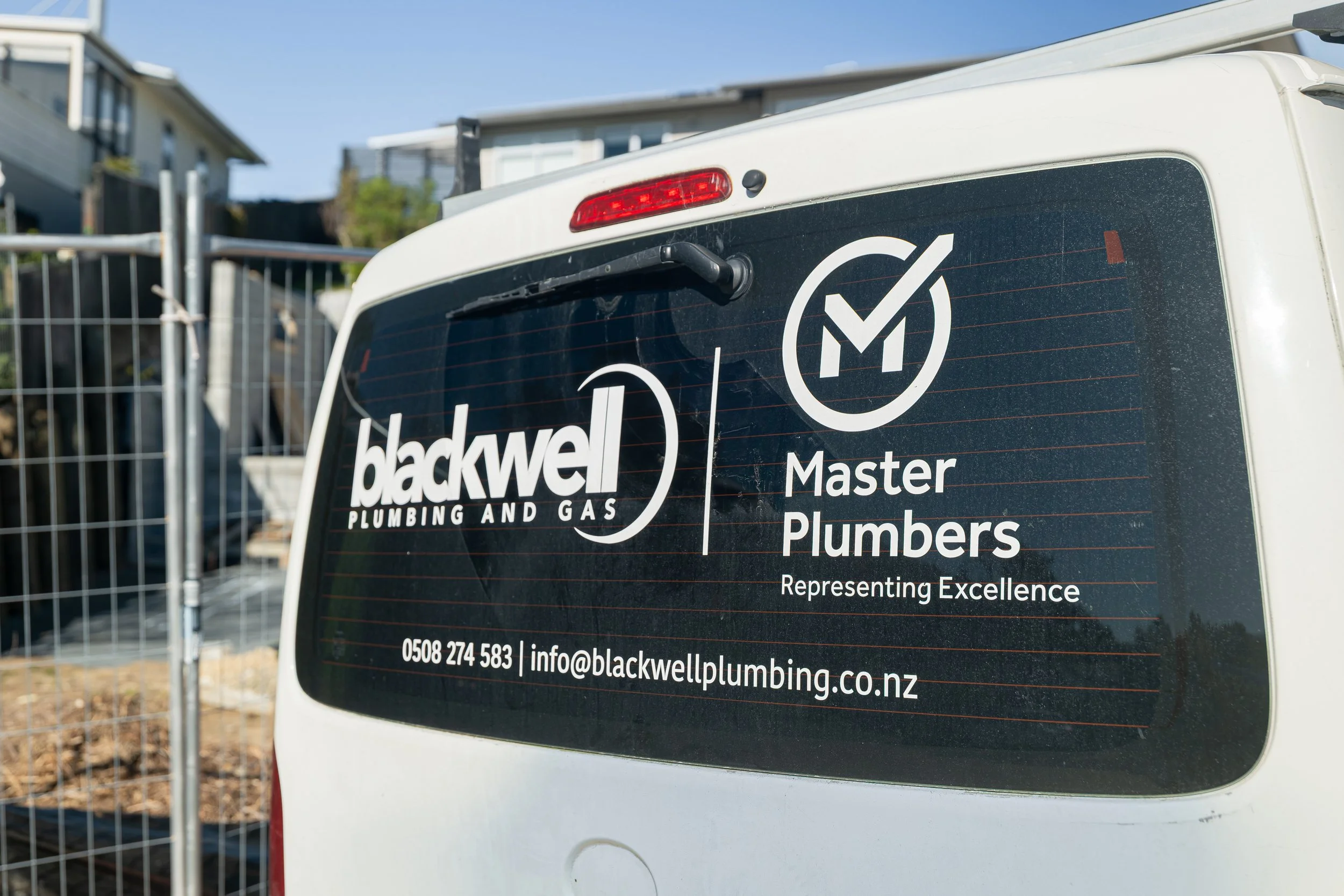 The rear window of a white van with black and white branding for Blackwell Plumbing and Gas, including the Master Plumbers logo, contact phone number, and email address. There is a chain-link fence and residential houses in the background.