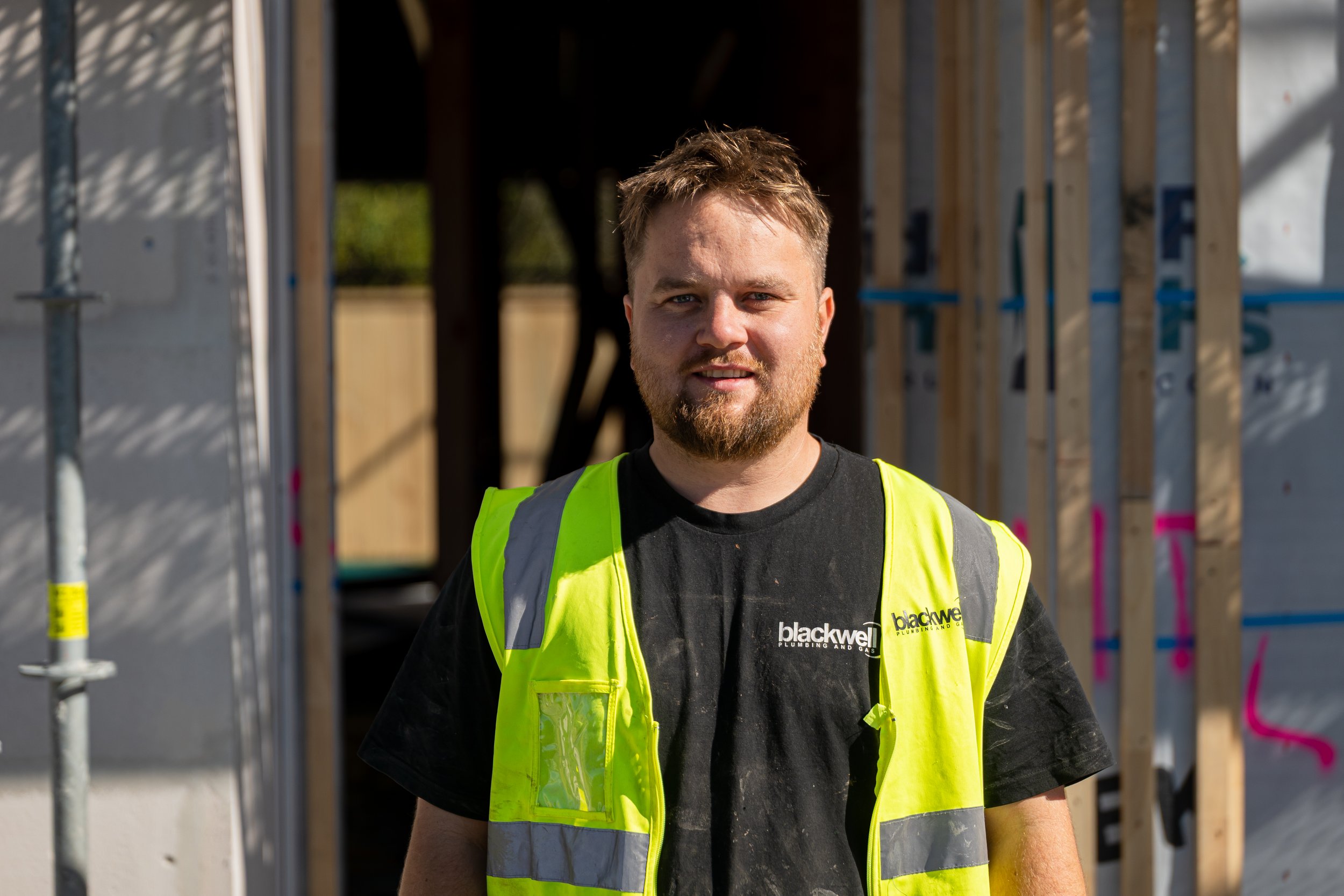 A construction worker wearing a black t-shirt with a company logo and a yellow safety vest standing on a construction site with wooden framing in the background.