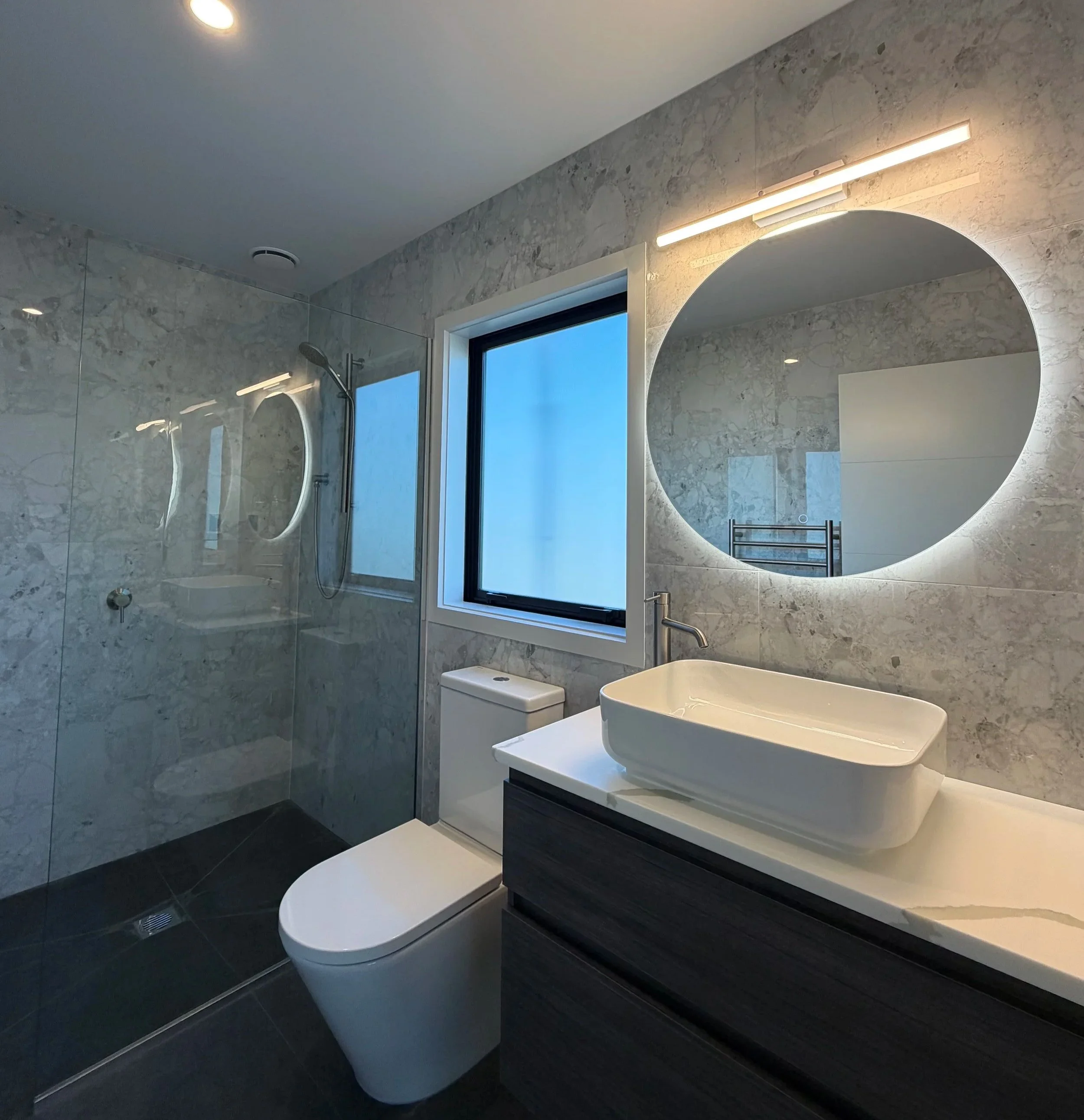 Modern bathroom with marble walls, a round mirror with LED lighting, a window, and a white toilet.