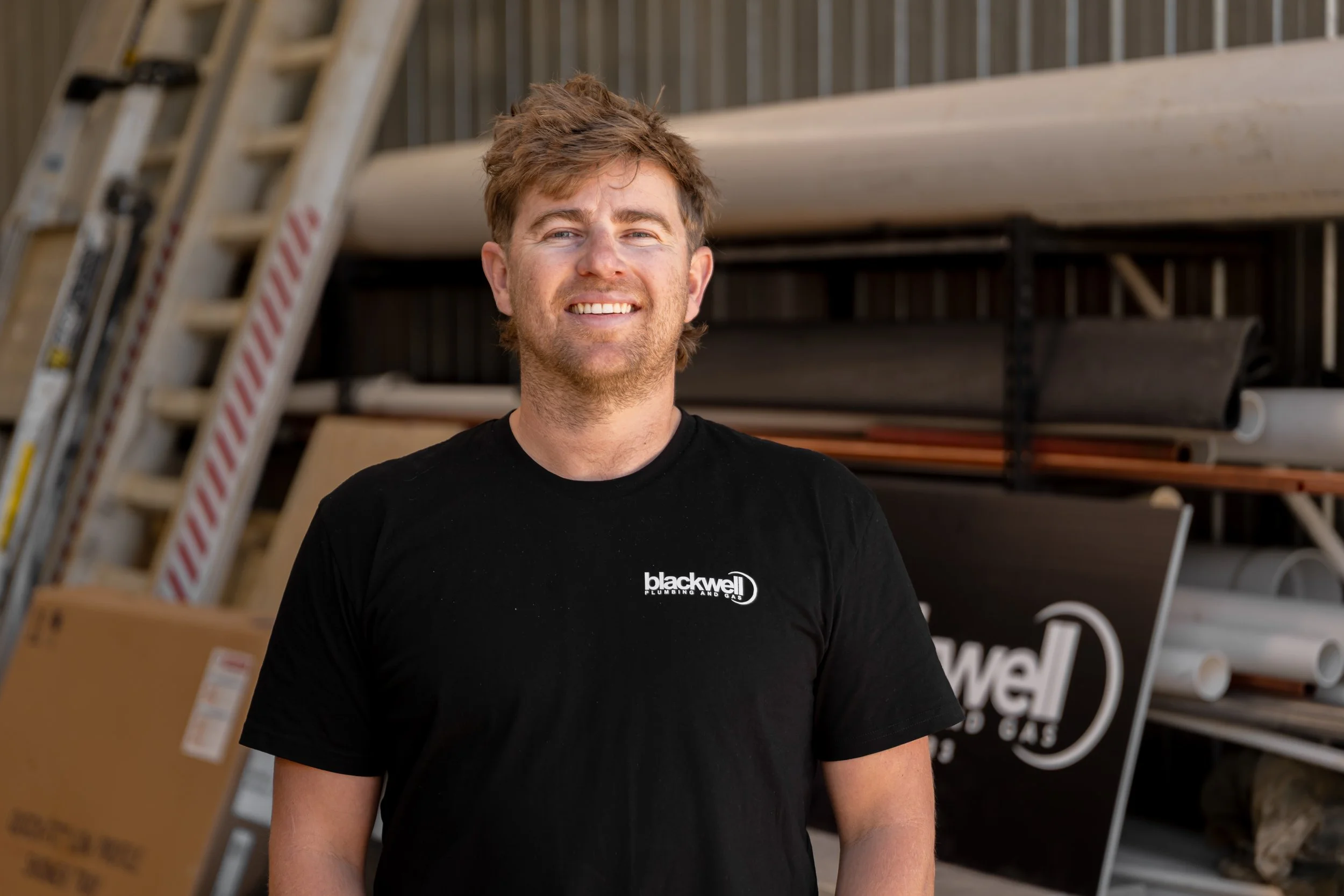 Young man smiling, wearing a black t-shirt with 'blackwell' logo, standing in hardware or plumbing store with pipes and ladders in background.