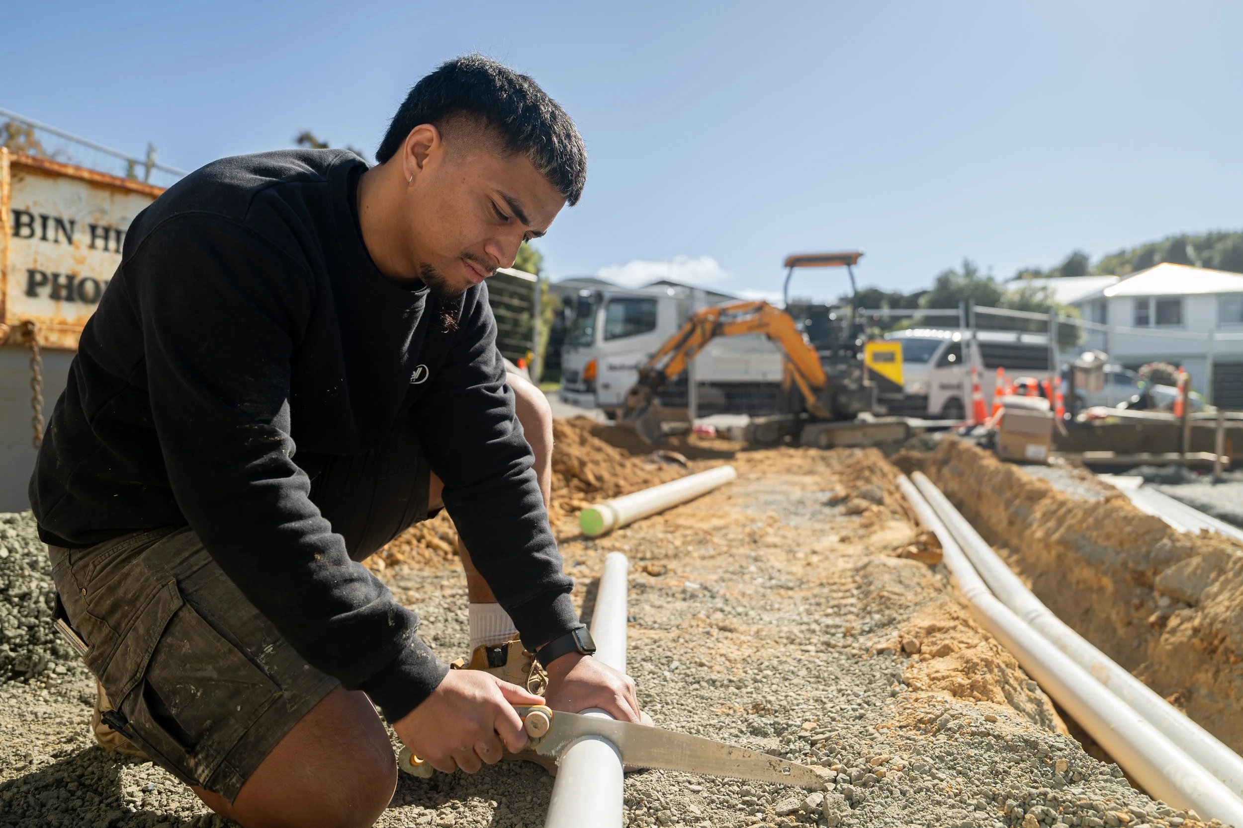 A man wearing a black hoodie and shorts is working on a construction site, cutting a pipe with a handsaw while kneeling on gravel.