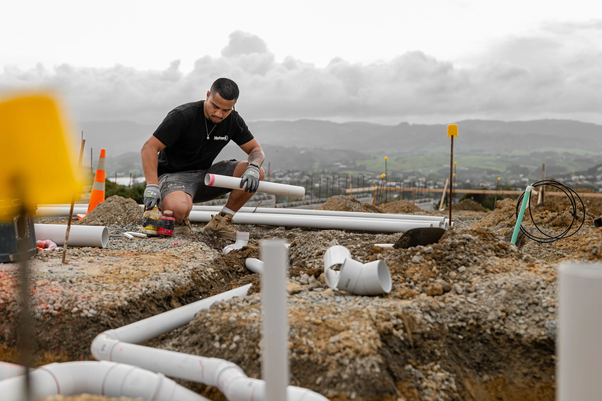 A plumber installing pipes on an outdoor site with cloudy skies and hills in the background.