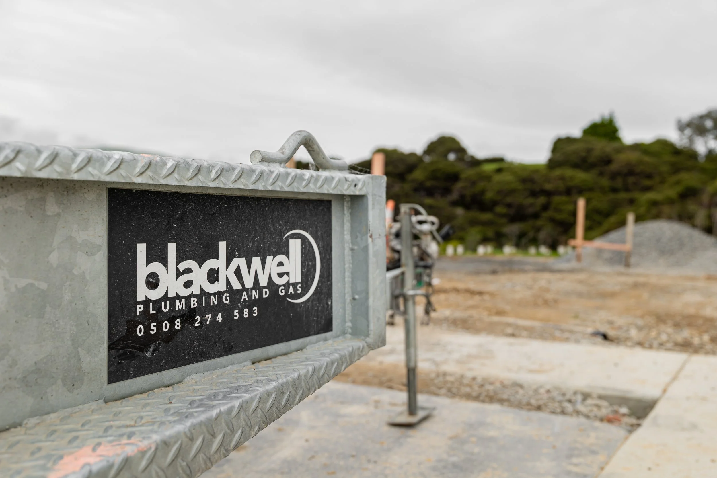Close-up of a metal sign with the logo and contact information for Blackwell Plumbing and Gas attached to a metal structure at a construction site. Blurred background of construction materials, scaffolding, and green trees under a cloudy sky.