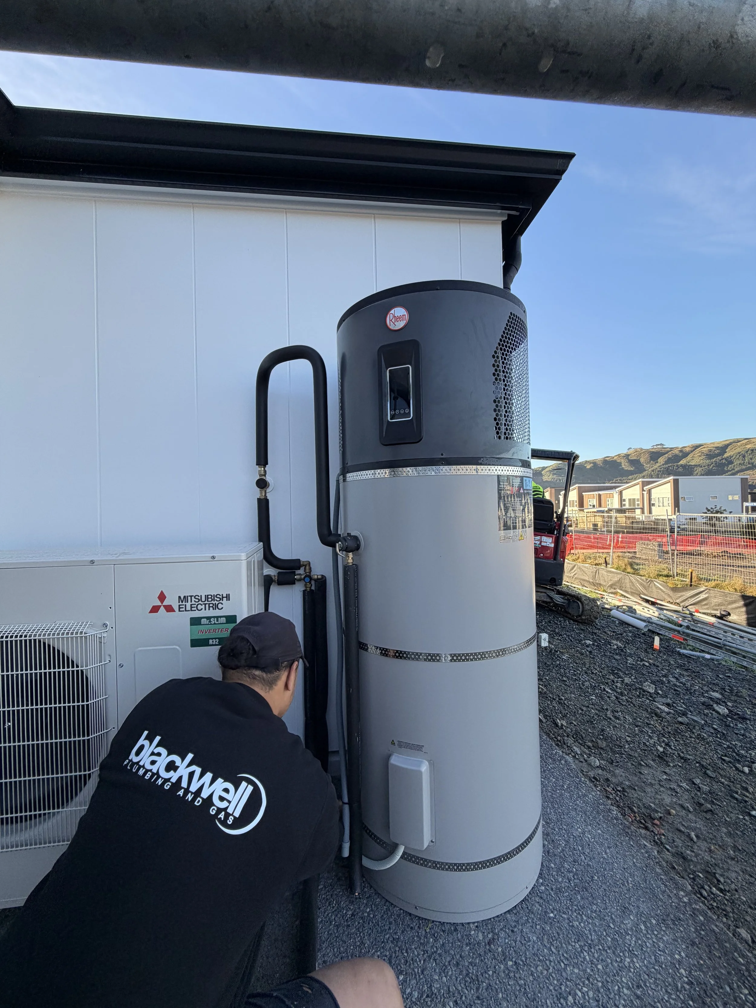 A man wearing a black Blackwell Plumbing hoodie is working on a large white cylindrical water heater with a black Rheem logo, outdoors near buildings and a chain-link fence, with a clear blue sky and hills in the background.