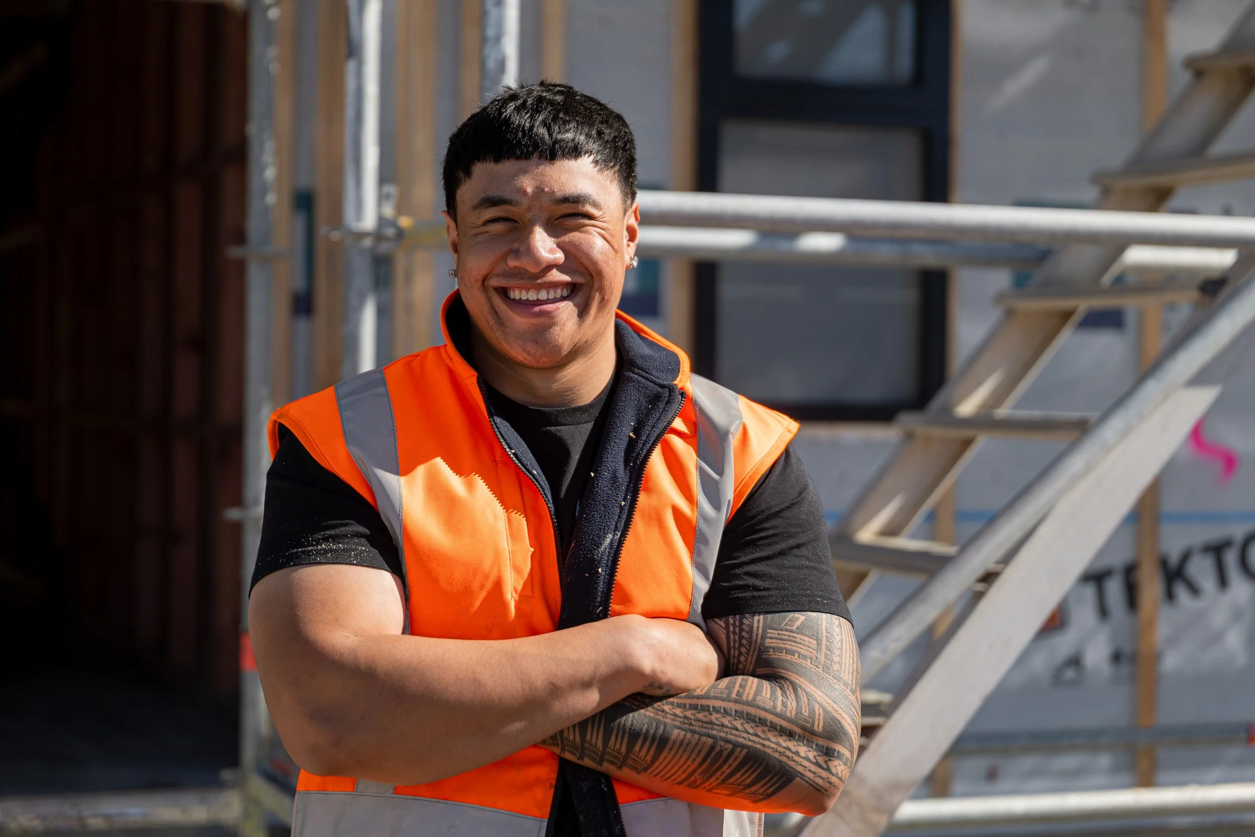 Smiling construction worker with tattoos wearing an orange safety vest and black shirt, standing outdoors near construction scaffolding.