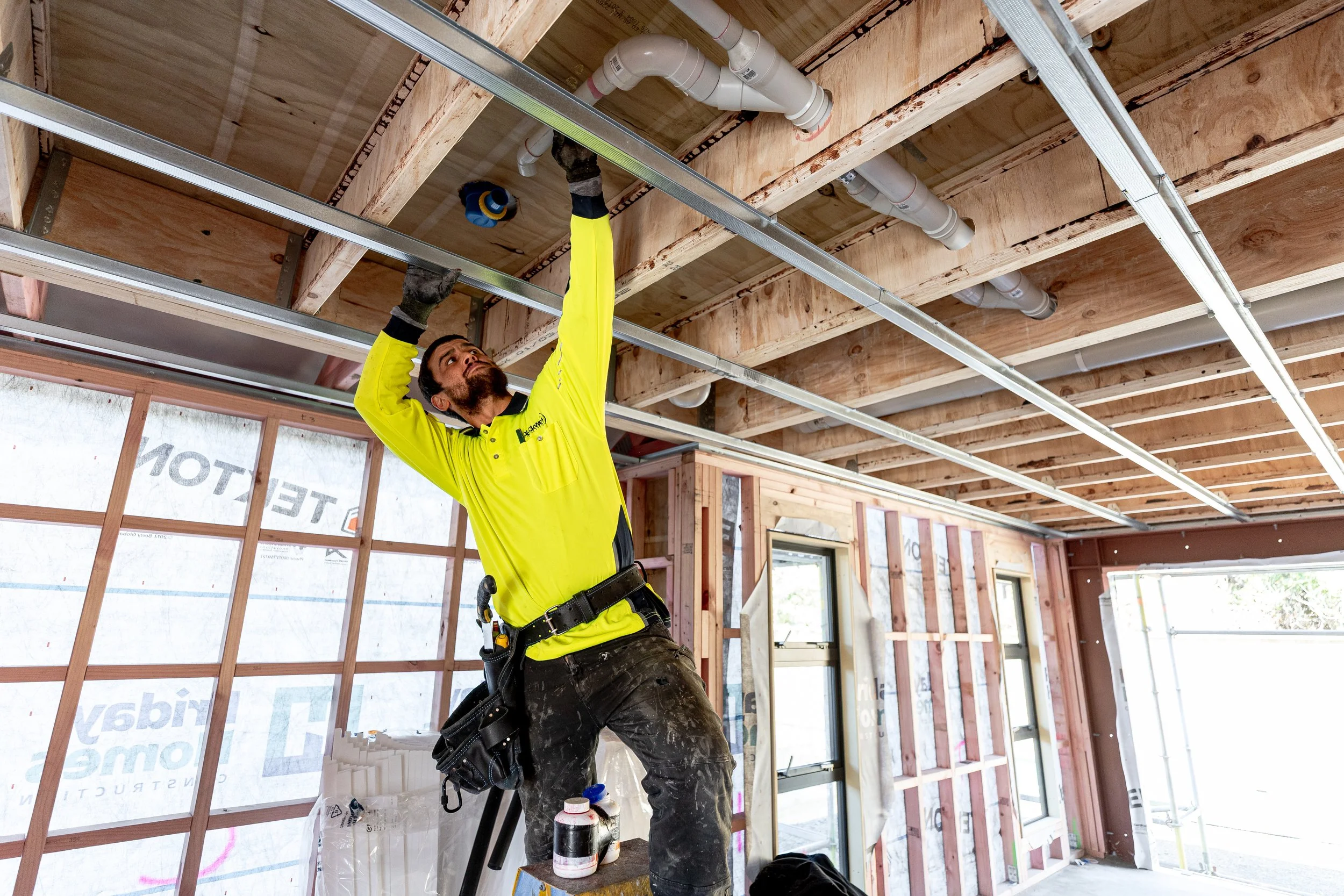 A construction worker wearing a bright yellow safety shirt and a tool belt, standing on a platform, working on the ceiling framework of a house under construction.