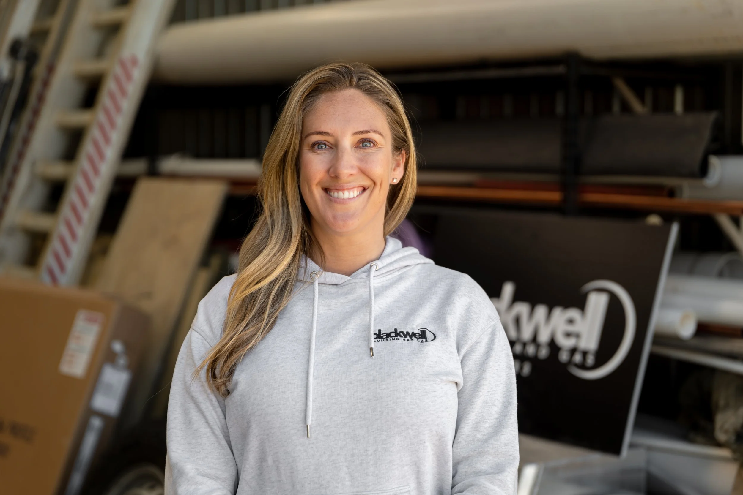 A smiling woman with long blonde hair wearing a light gray hoodie with a logo that reads 'Blackwell' standing in front of a shelving unit of building supplies, including pipes and a black sign with white text.