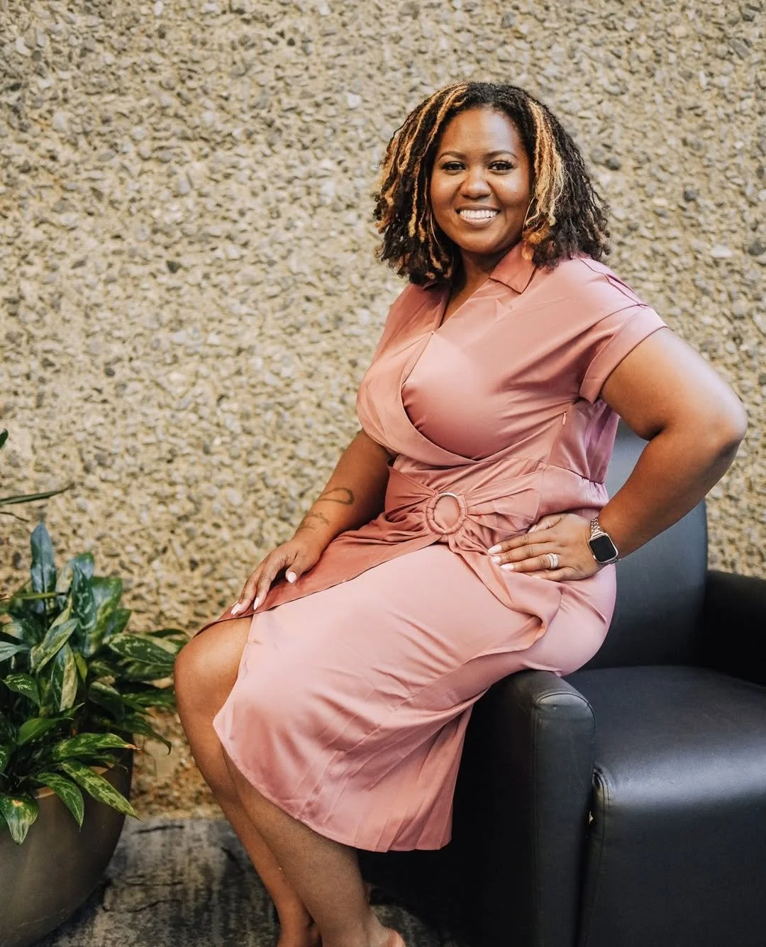 Woman with curly hair sitting on a black chair, wearing a pink dress, smiling, with a textured beige wall and plant in the background.