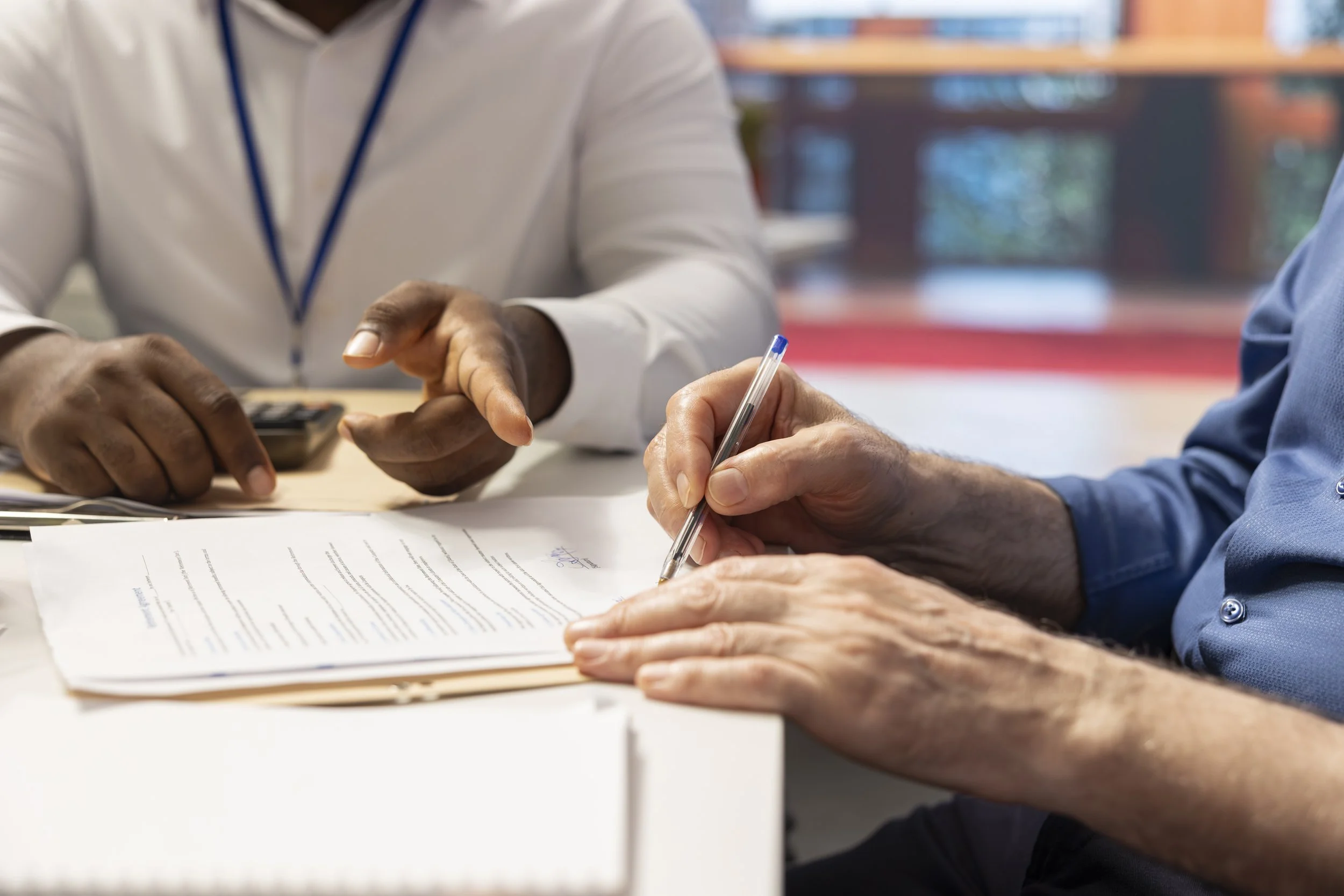 Two people sitting at a table; one is writing on a document with a pen, the other is gesturing with their hands near the document, which appears to be paper or a form.